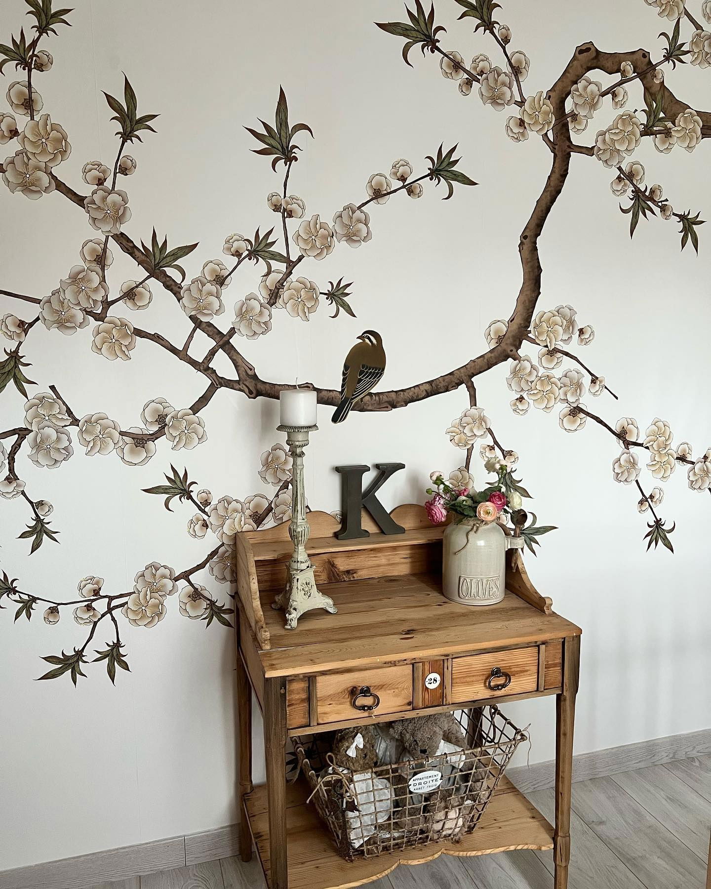 Rustiek houten bureau met bloemenbehang op de achtergrond. Vogel op tak, kaars en "Olives" vaas erop. Draadmand met knuffels eronder.