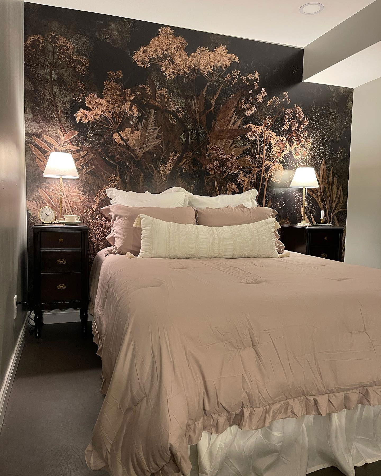 Schlafzimmer mit floraler Tapete, Lampen auf dunklen Holz Nachttischen und einem Bett mit lilafarbener Bettwäsche.