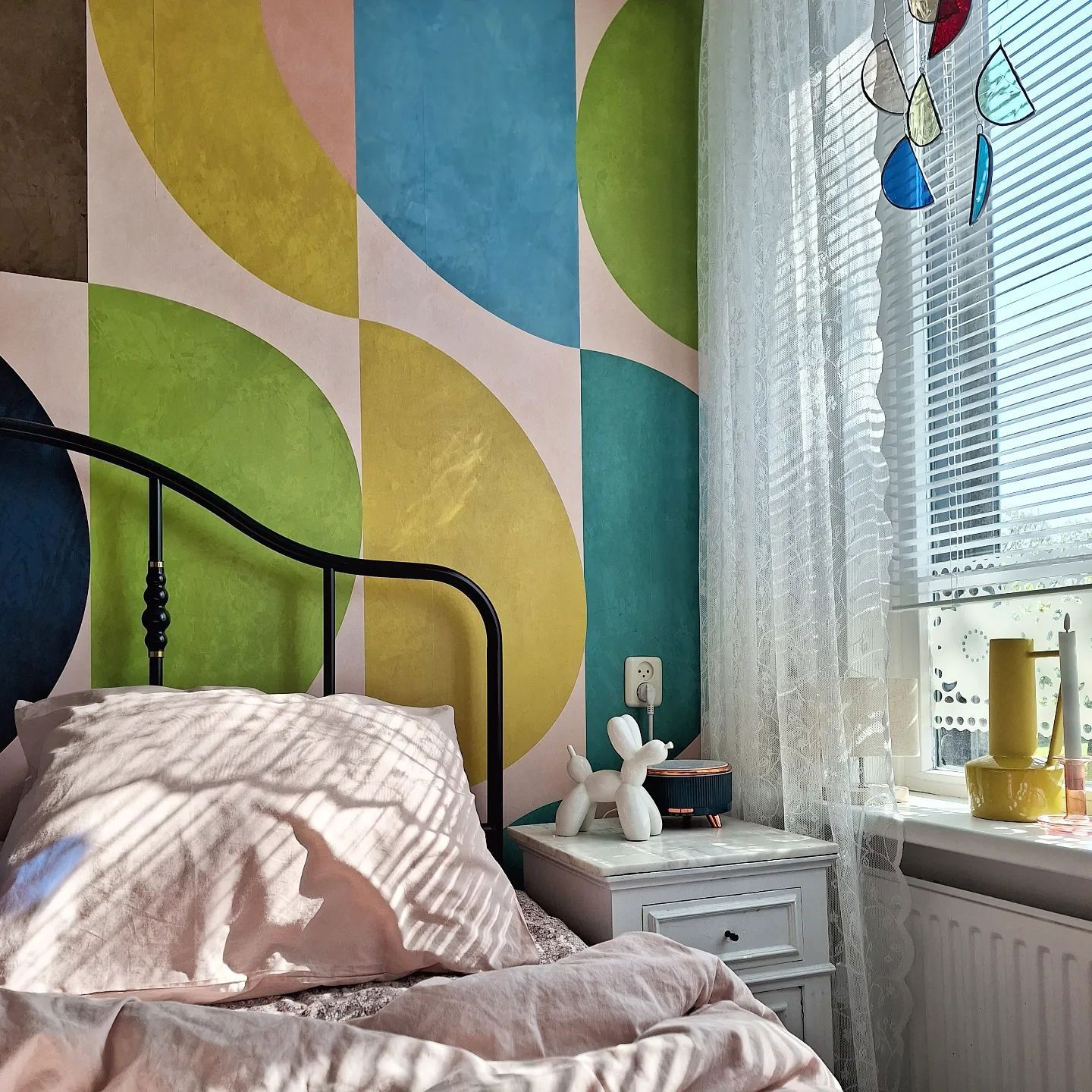 Bedroom with black metal bed frame, pink bedding, and colorful geometric wallpaper. Sunlight streams through window with sheer curtains.