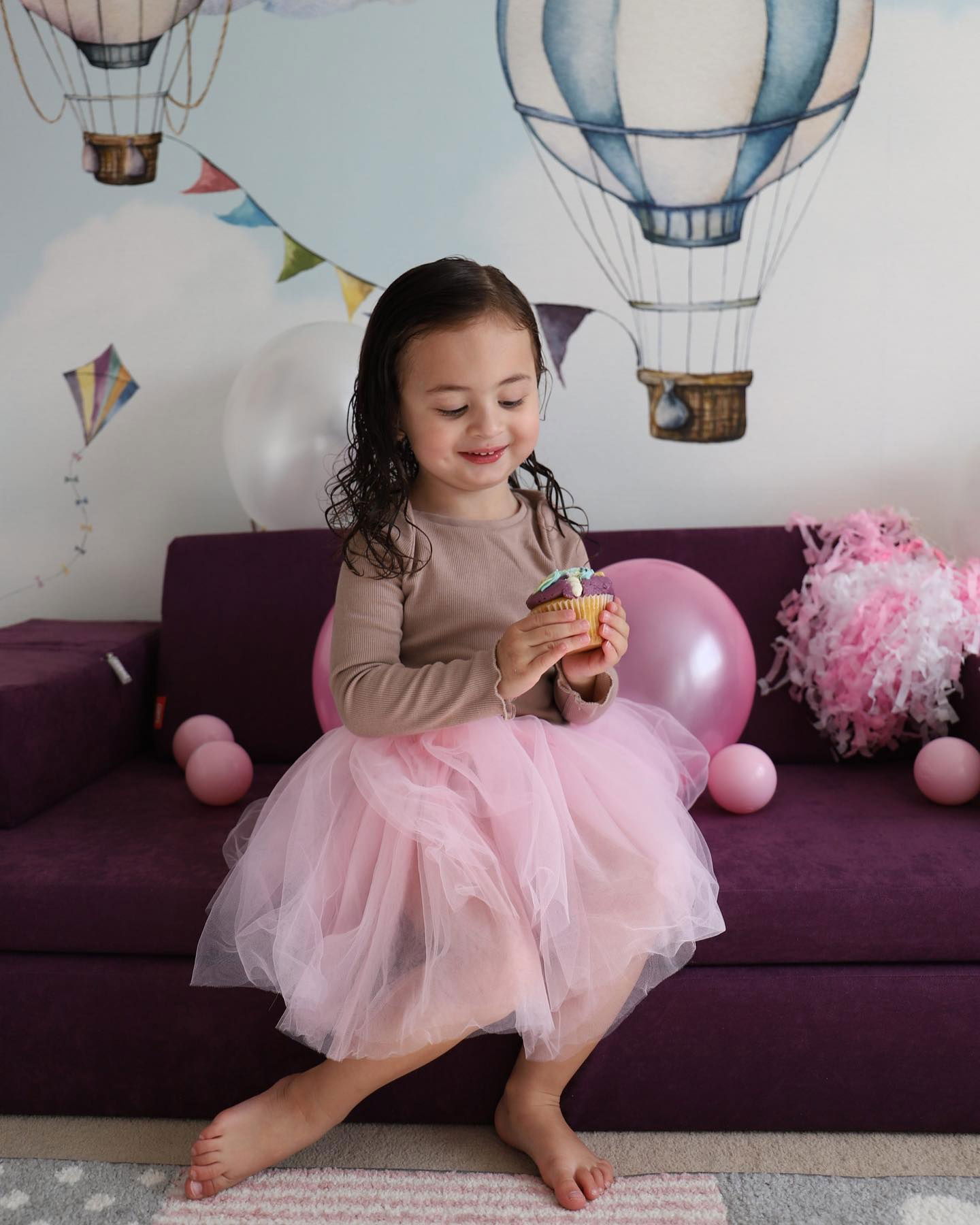 Une jeune fille en tutu rose est assise sur un canapé violet, tenant un cupcake. Des ballons et des décorations l'entourent.