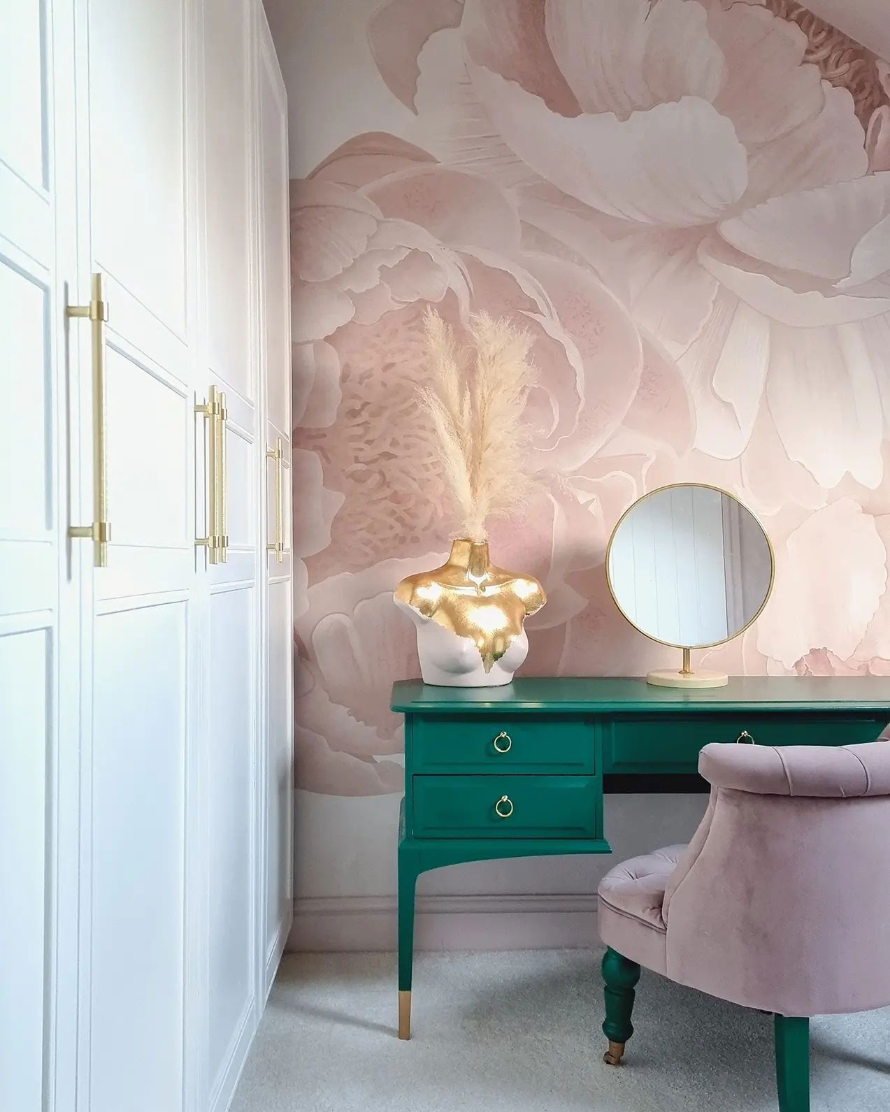 Scena di una camera da letto con mobile da bagno verde, sedia rosa e carta da parati floreale. Un vaso a busto contiene erba di pampa.