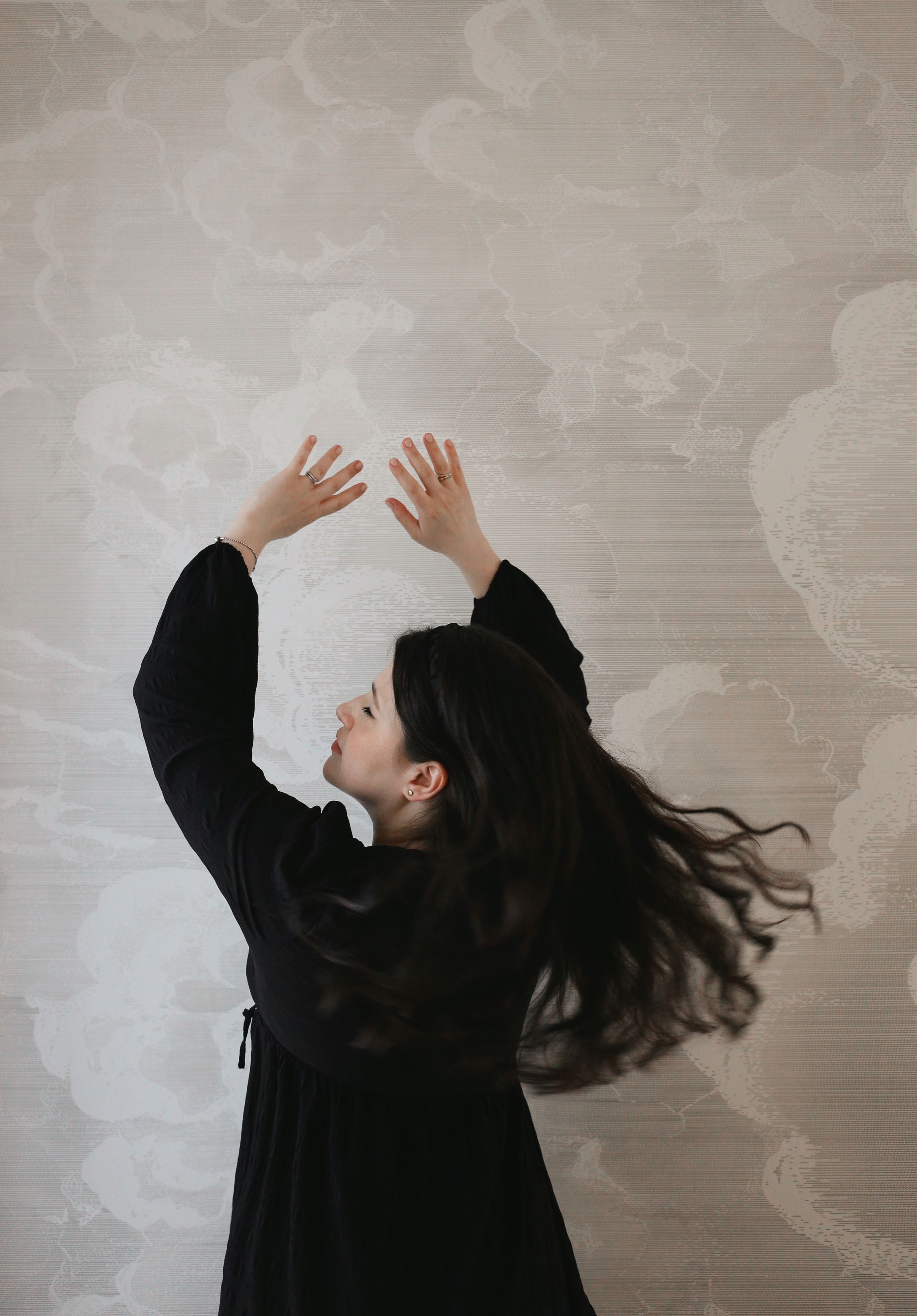 Woman in black dress with arms raised, hair flowing against cloud-patterned wall.