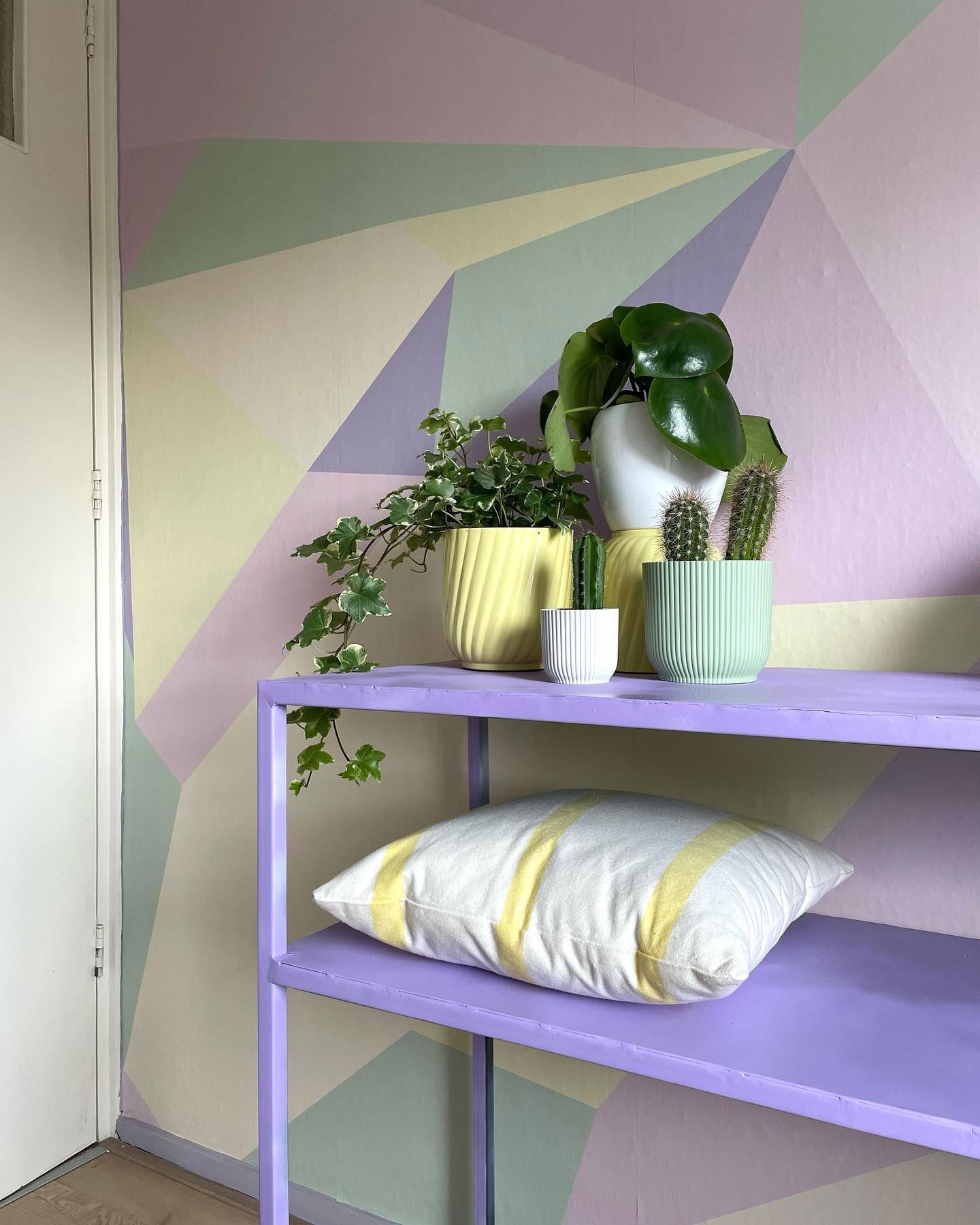 Pastel geometric wall with purple shelf holding plants and a striped pillow.
