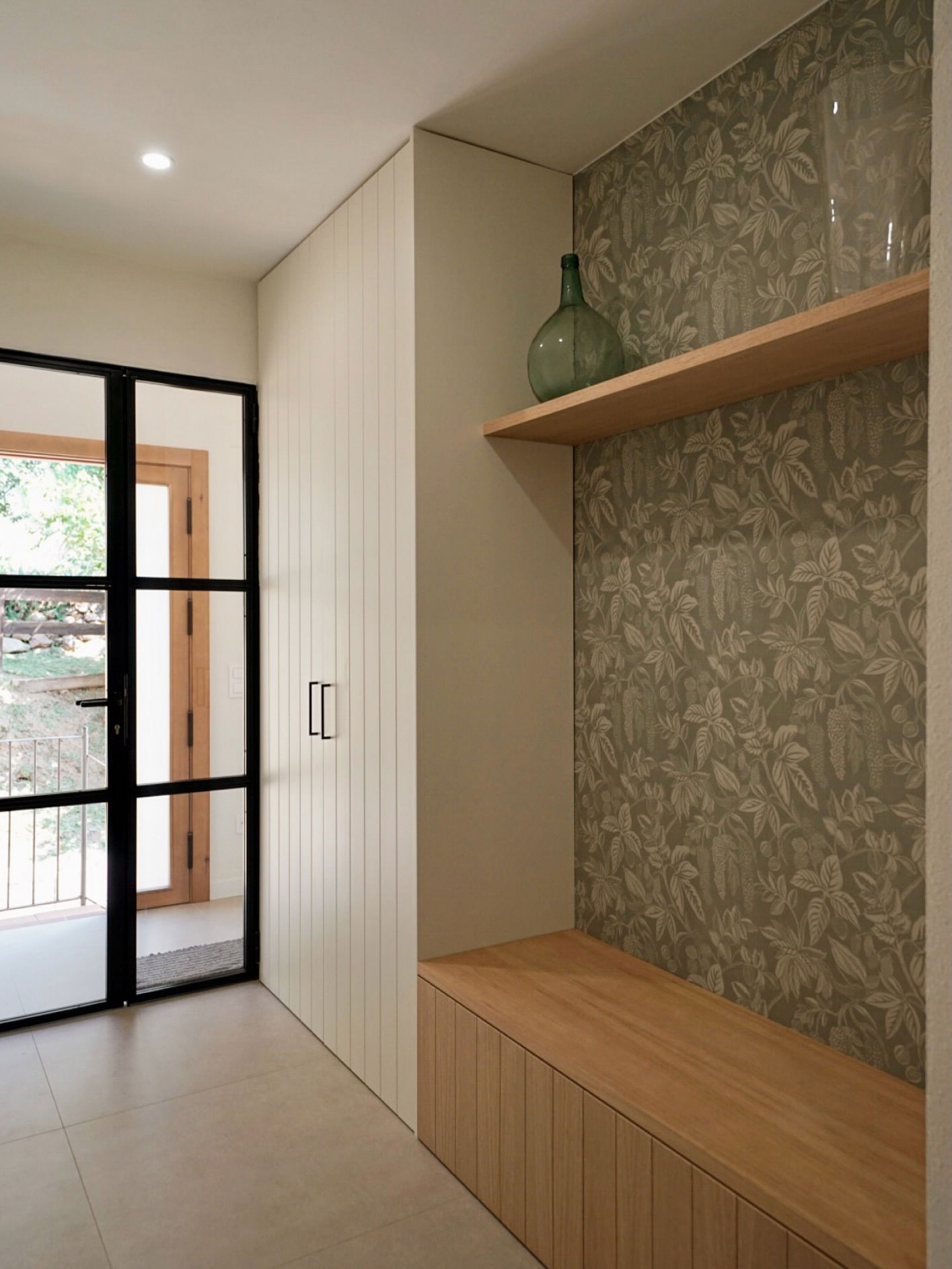 Hallway with black-framed glass door, white closet, and wooden bench with leaf-patterned wallpaper.