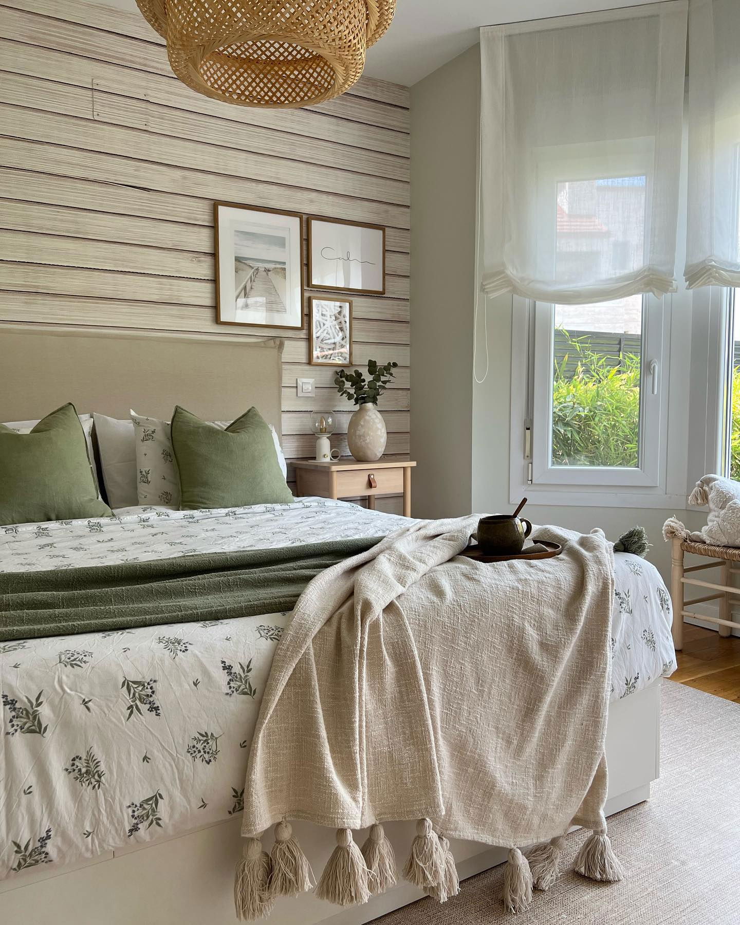 Cozy bedroom with floral bedding, green pillows, and a tasseled throw blanket. Woven pendant light and framed art above the bed.
