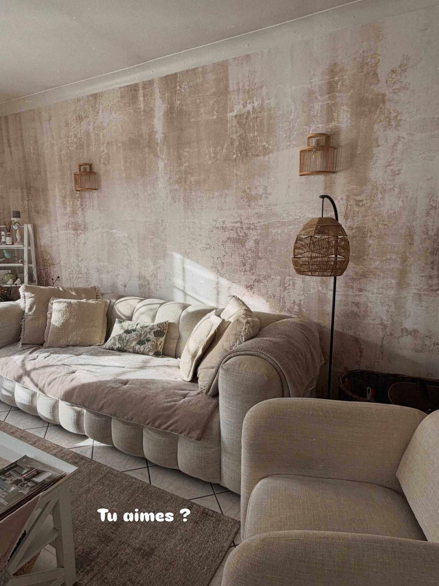 En beige tuftet sofa med pyntepuder og tæpper i et rum med teksturerede vægge og naturligt lys.