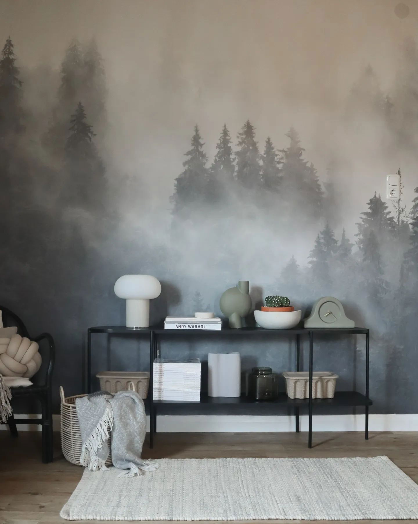Interior with forest mural, black shelving unit, and woven rug. Décor includes a lamp, books, vases, and a clock.