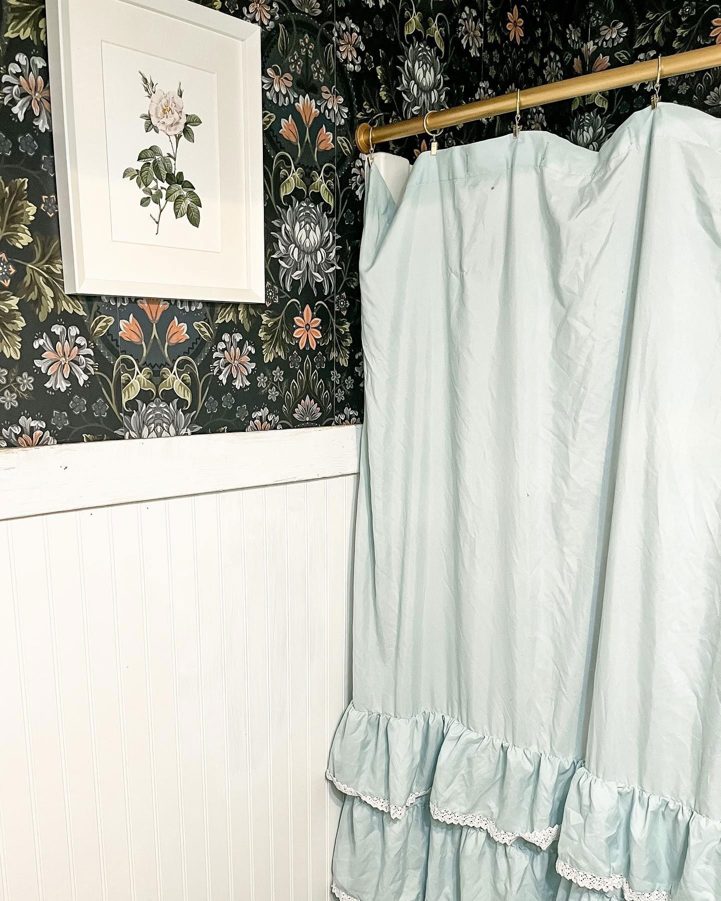 Badkamer met bloemenbehang, witte lambrisering, ingelijste bloemenkunst en lichtblauwe douchegordijn met ruches.