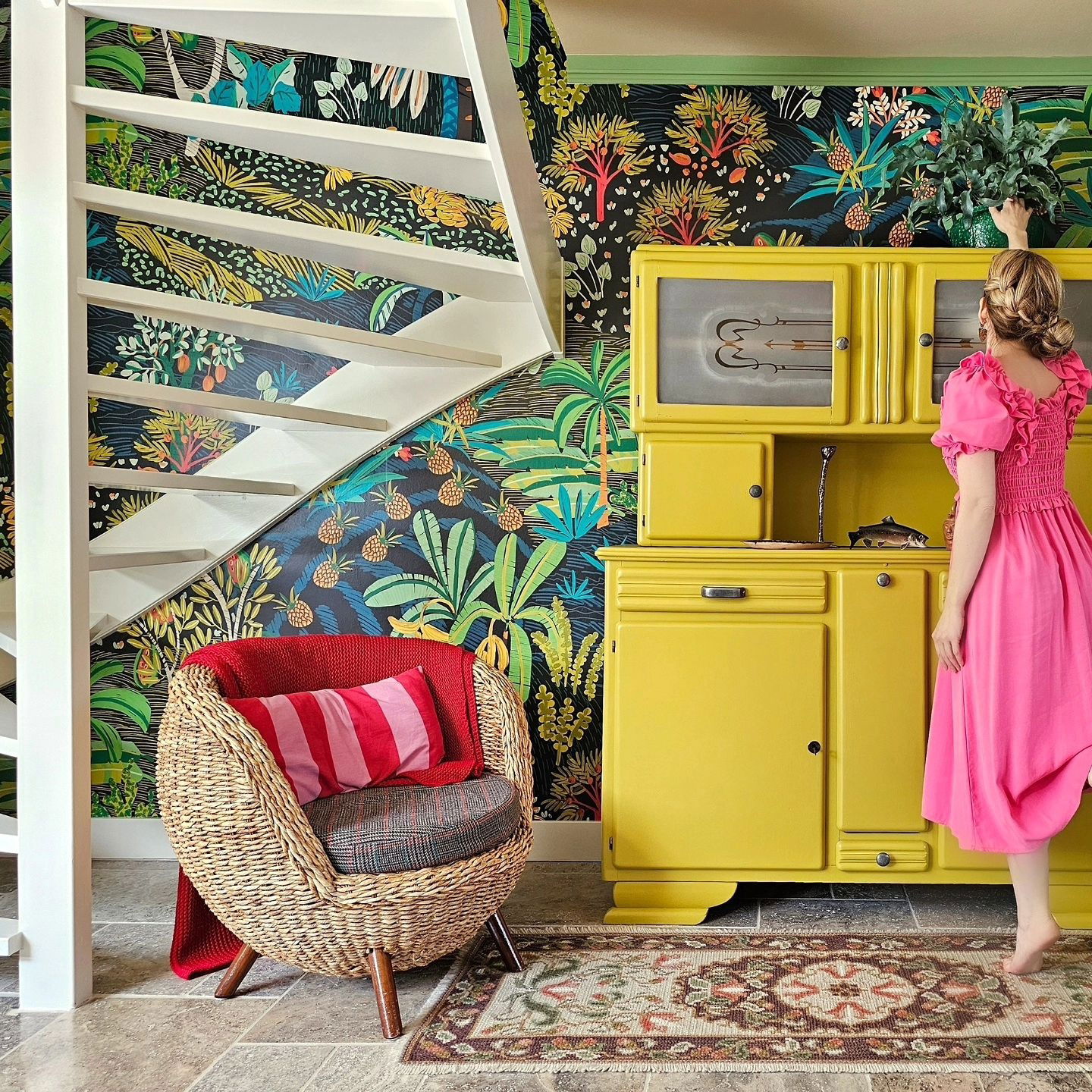 Eclectic room with tropical wallpaper, yellow hutch, and wicker chair. A woman in a pink dress arranges flowers on the hutch.