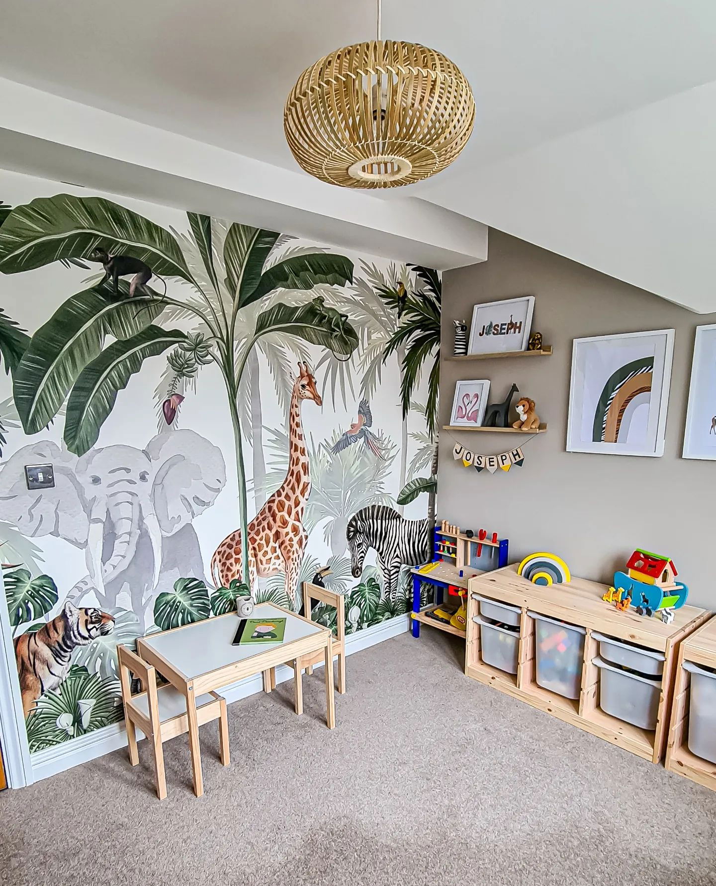 Child's room with jungle mural, wooden table and chairs, and toy storage. Wicker light fixture hangs from the ceiling.