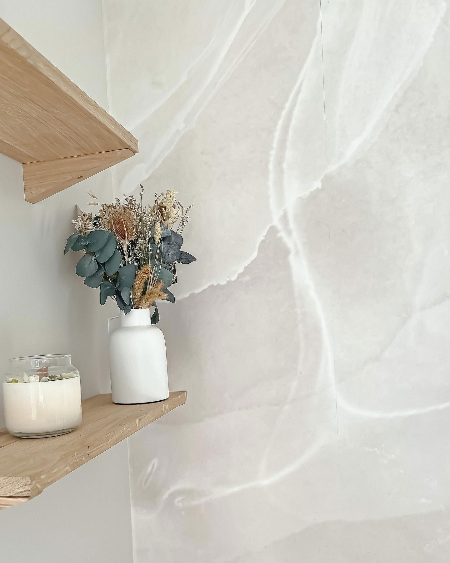 Natura morta su scaffali di legno: candela, composizione di fiori secchi in vaso bianco, parete con motivo marmorizzato.