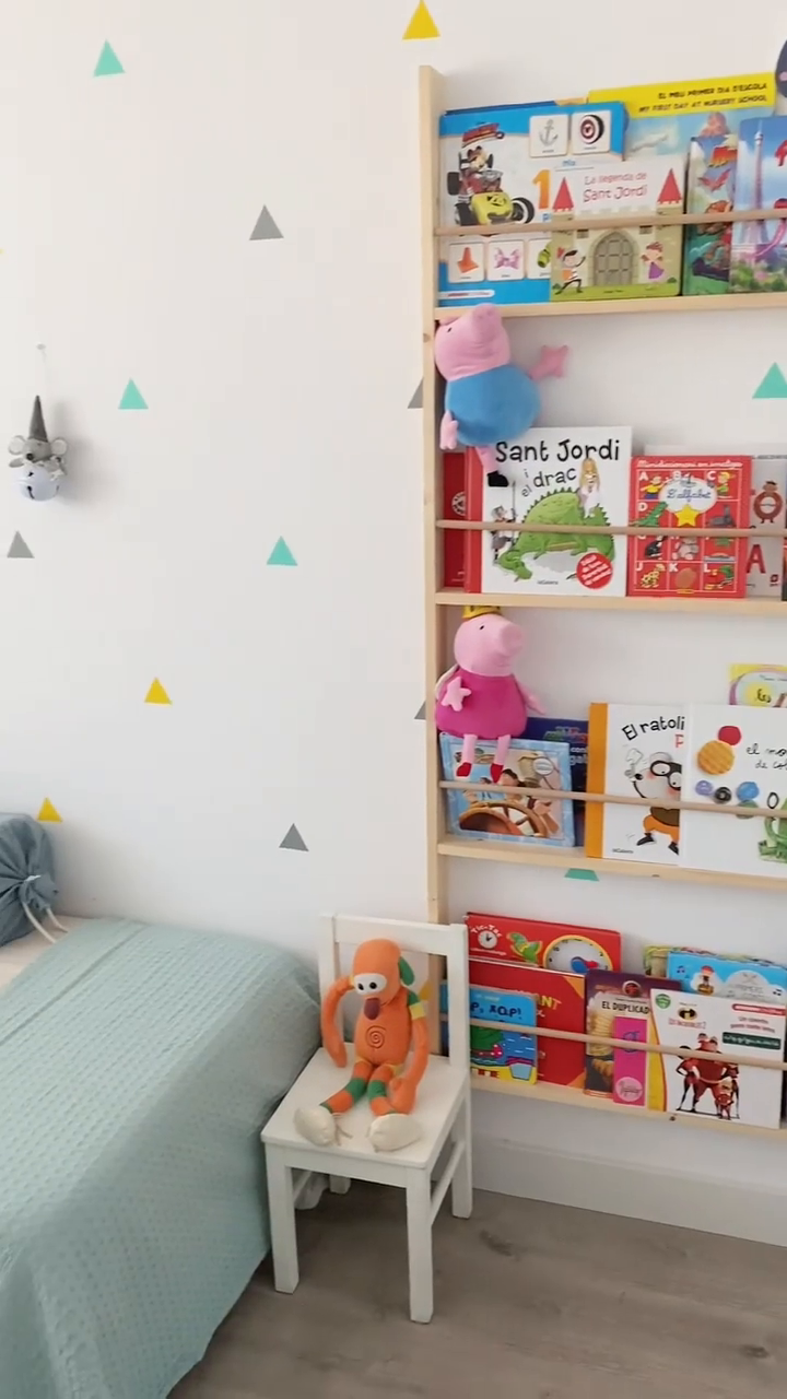 Child's room with light blue bedspread, triangle wall decals, bookshelf with books and stuffed animals, and a toy on a white chair.