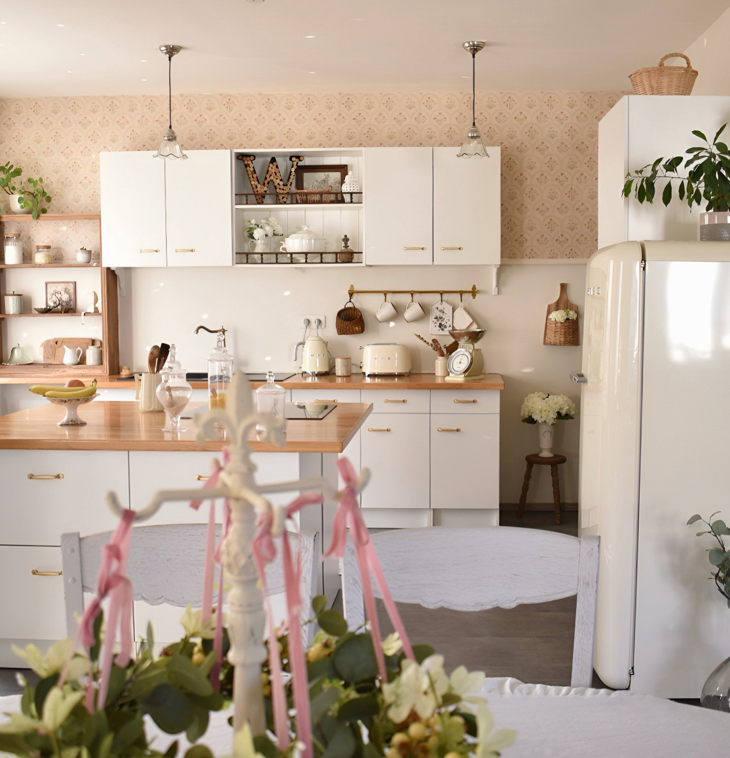Vitt kök, träbänkskivor, vita skåp, beige tapet. Vit kyl till höger. Bord i förgrunden med rosa band som mittpunkt.