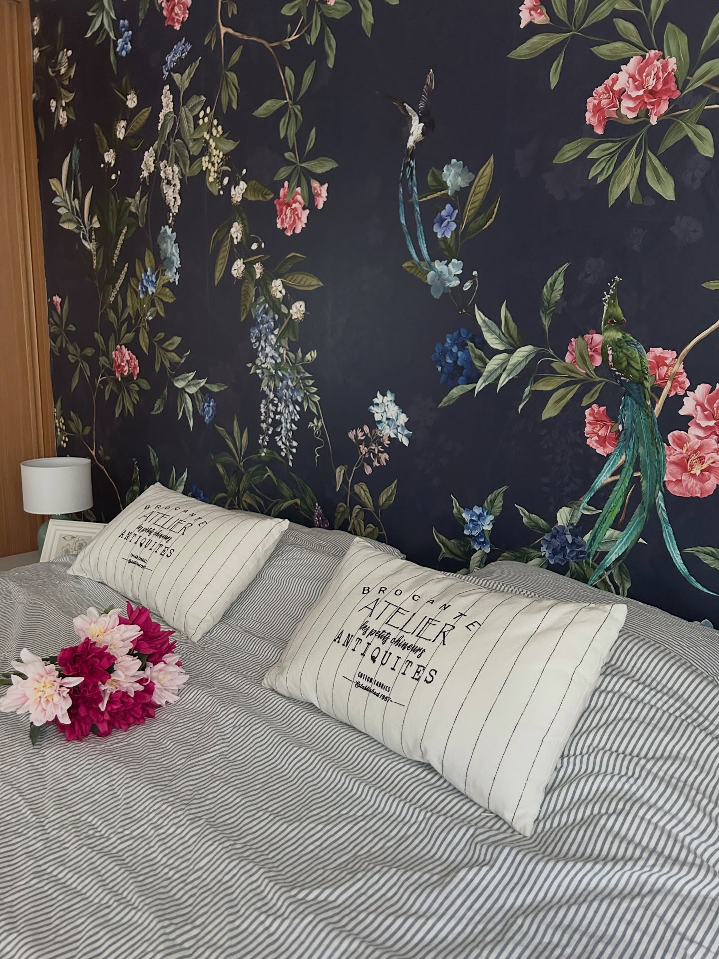 Bedroom with dark floral wallpaper, striped bedding, two decorative pillows, and a bouquet of pink flowers.