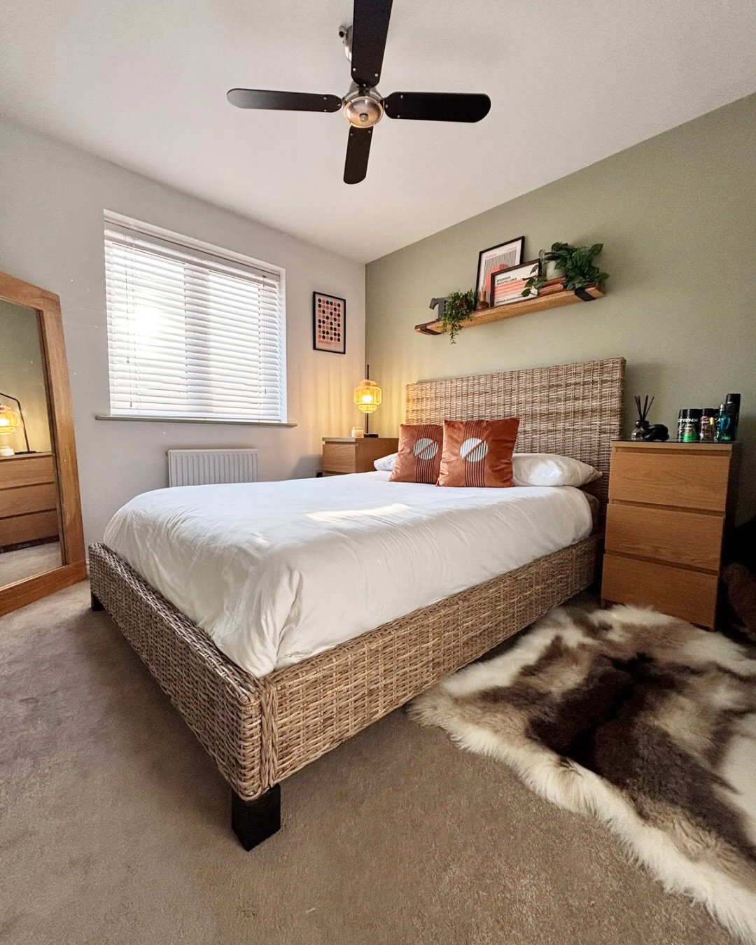 Ein Schlafzimmer mit einem Webbett, einer weißen Bettdecke und orangen Kissen. Eine grüne Wand hat ein Regal mit Pflanzen und Kunst. Ein Teppich aus Kunstpelz liegt auf dem Boden.