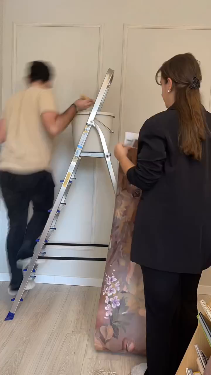 Two people hanging floral wallpaper. One person is on a ladder with a bucket.
