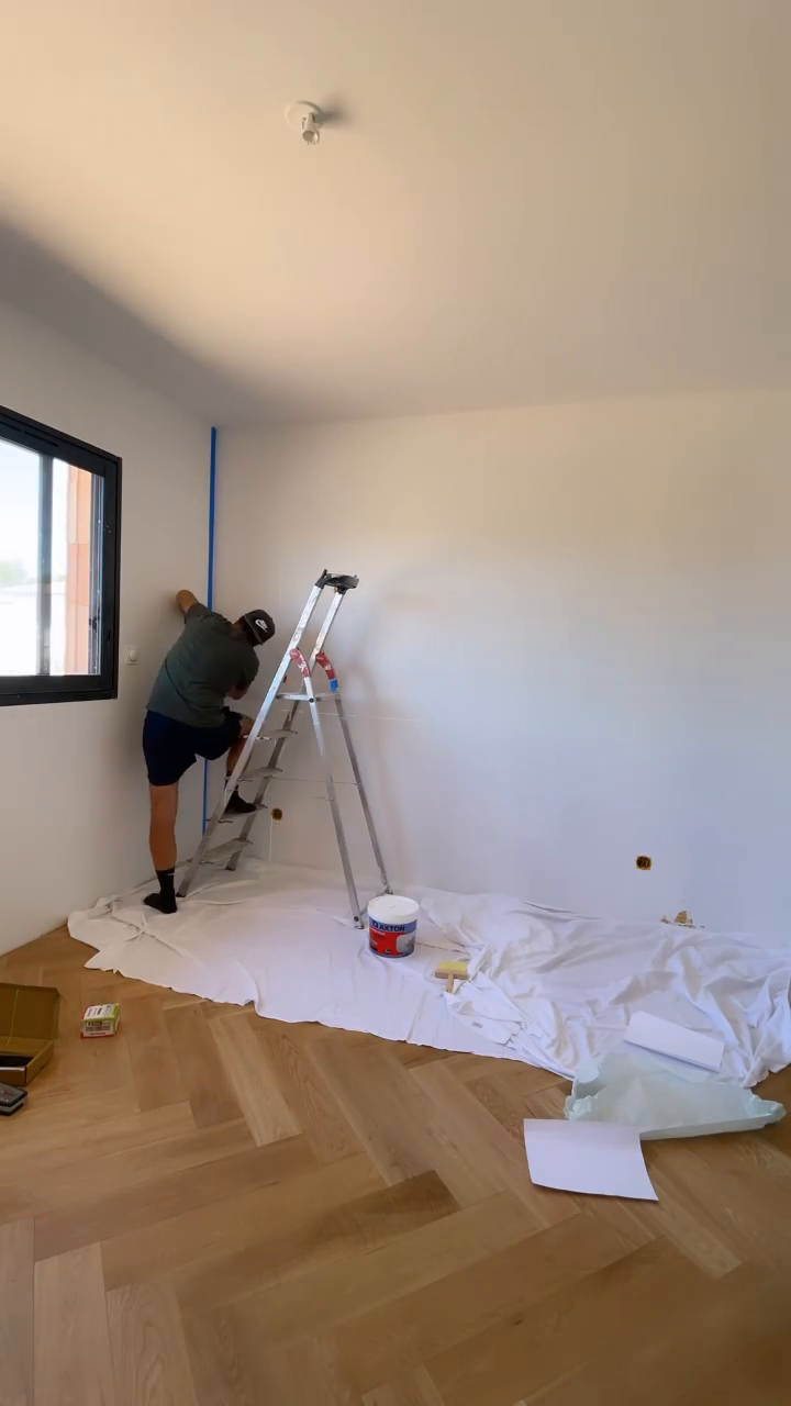 A person on a ladder tapes a wall in a room with wood floors. A white tarp covers the floor. A can of Axton paint is visible.