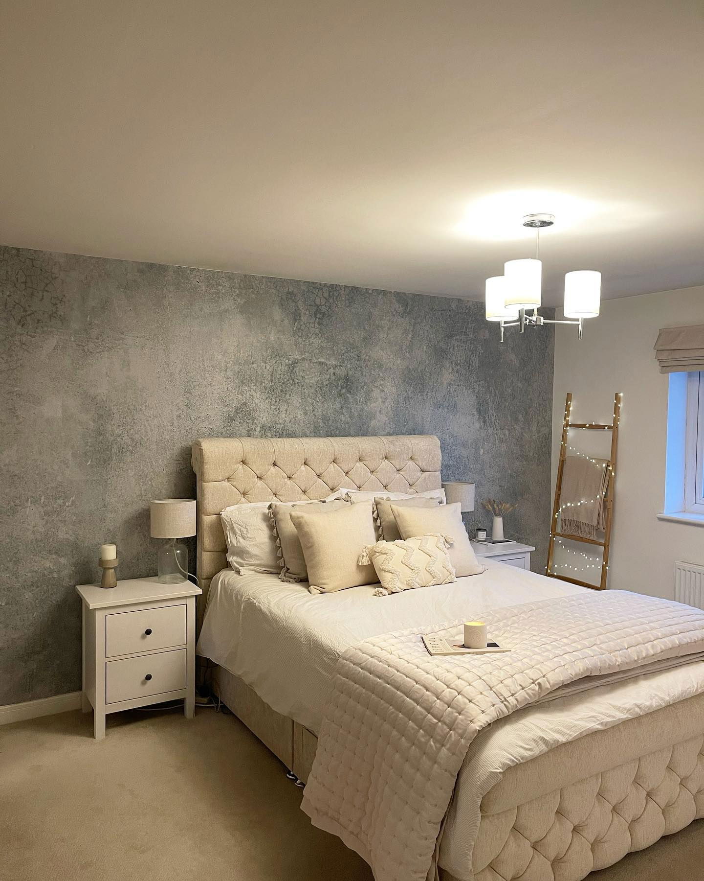 Bedroom with tufted beige bed, textured gray accent wall, and ladder with fairy lights.