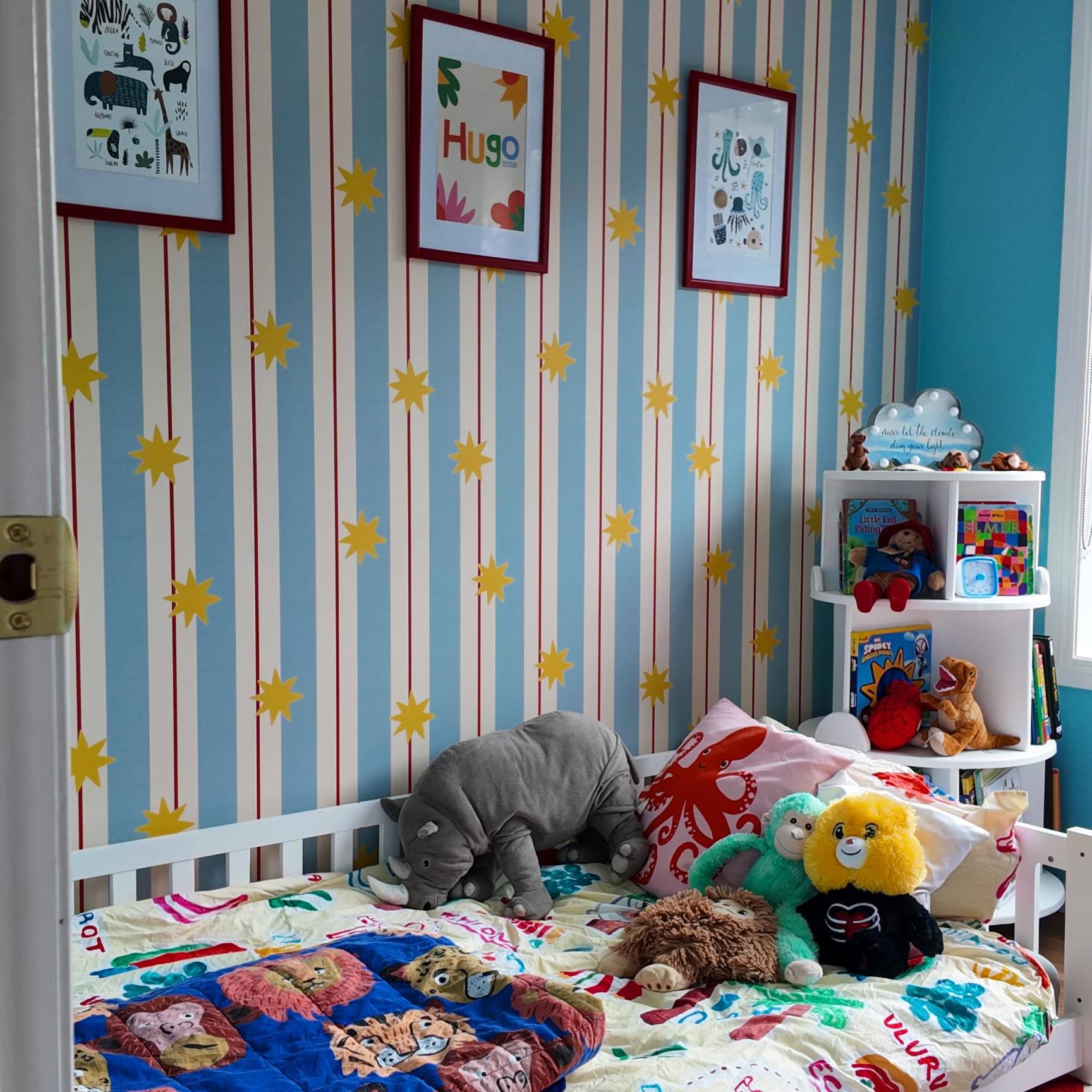 Fondo de pantalla a rayas azules y blancas con estrellas amarillas. Una cuna blanca tiene mantas de colores y animales de peluche. Una estantería blanca tiene libros y juguetes.