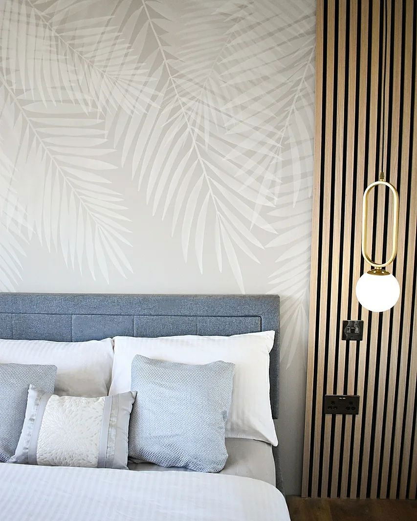 Dormitorio con cabecero gris, ropa de cama blanca y papel tapiz de hojas de palma. Una pared de listones de madera cuenta con una lámpara colgante dorada.
