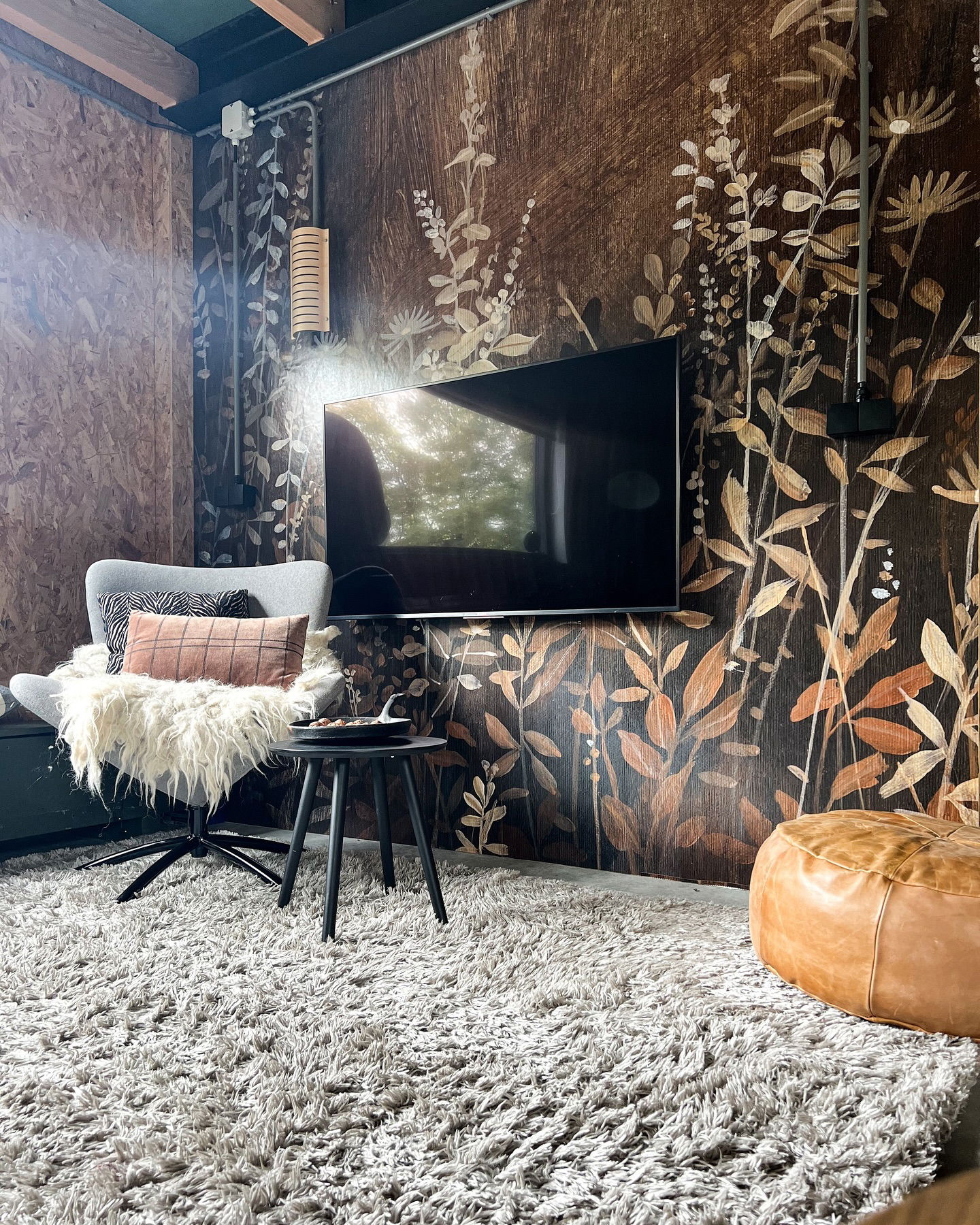 Cozy living room with floral wallpaper, gray chair, shag rug, and leather pouf.