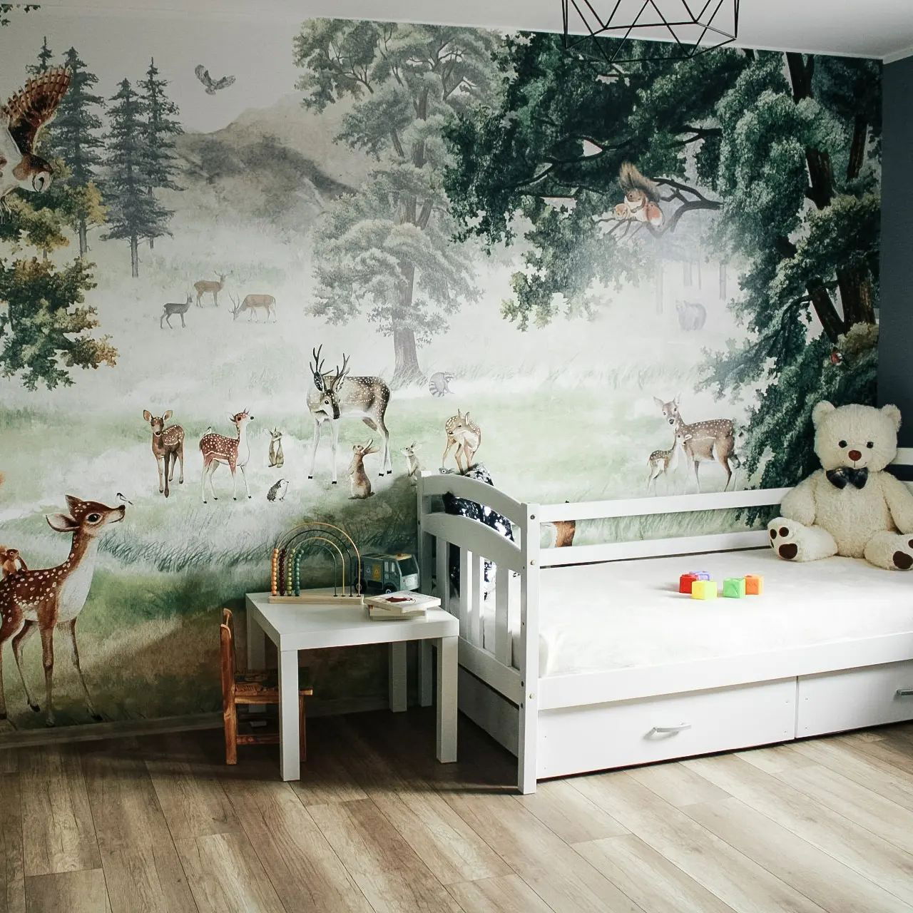 Chambre d'enfant avec une murale de forêt, un lit blanc avec un ours en peluche et une petite table blanche avec une chaise.