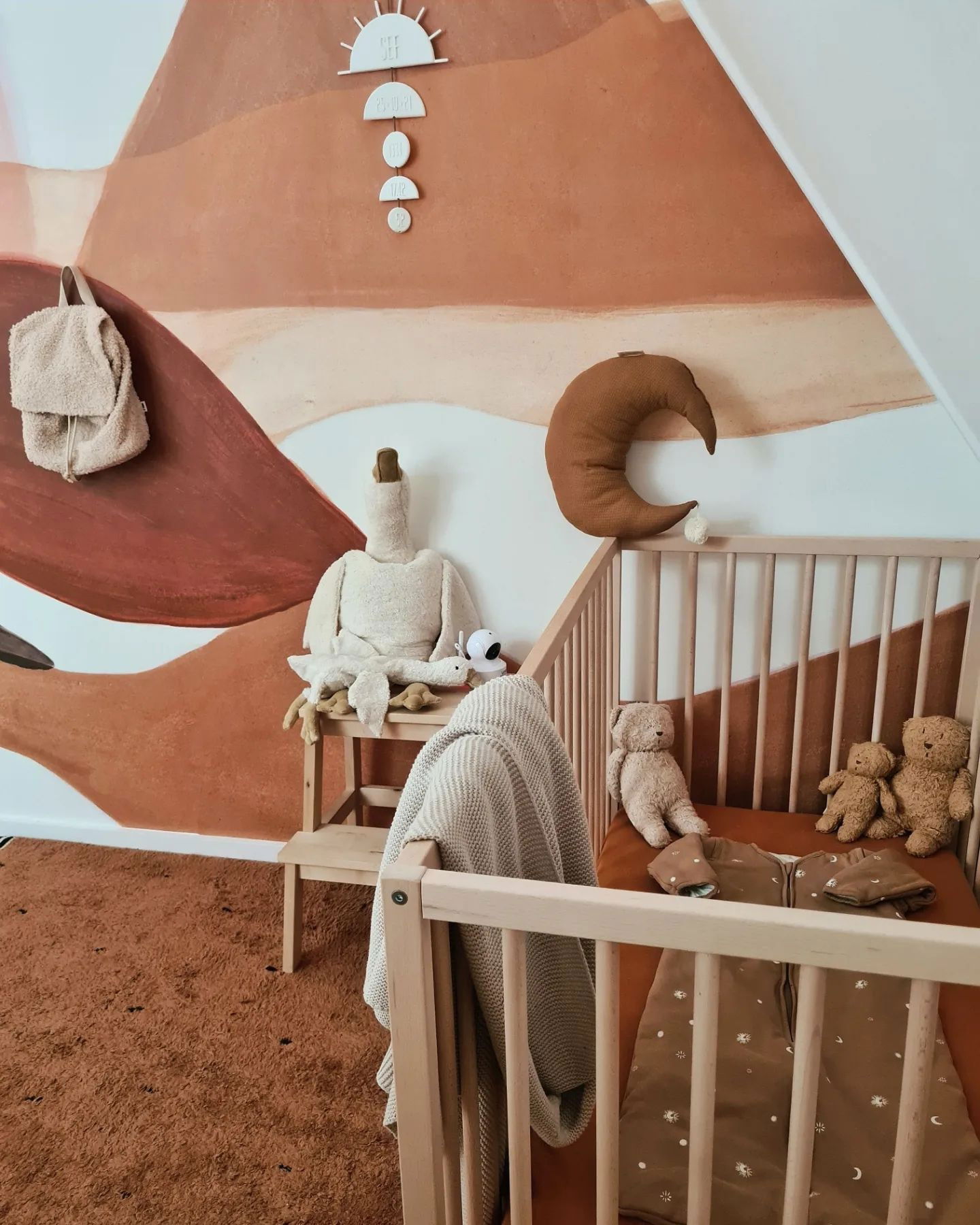 Nursery with crib, stuffed animals, and earth-tone mural. Moon pillow hangs above crib with teddy bears and sleeping bag inside.