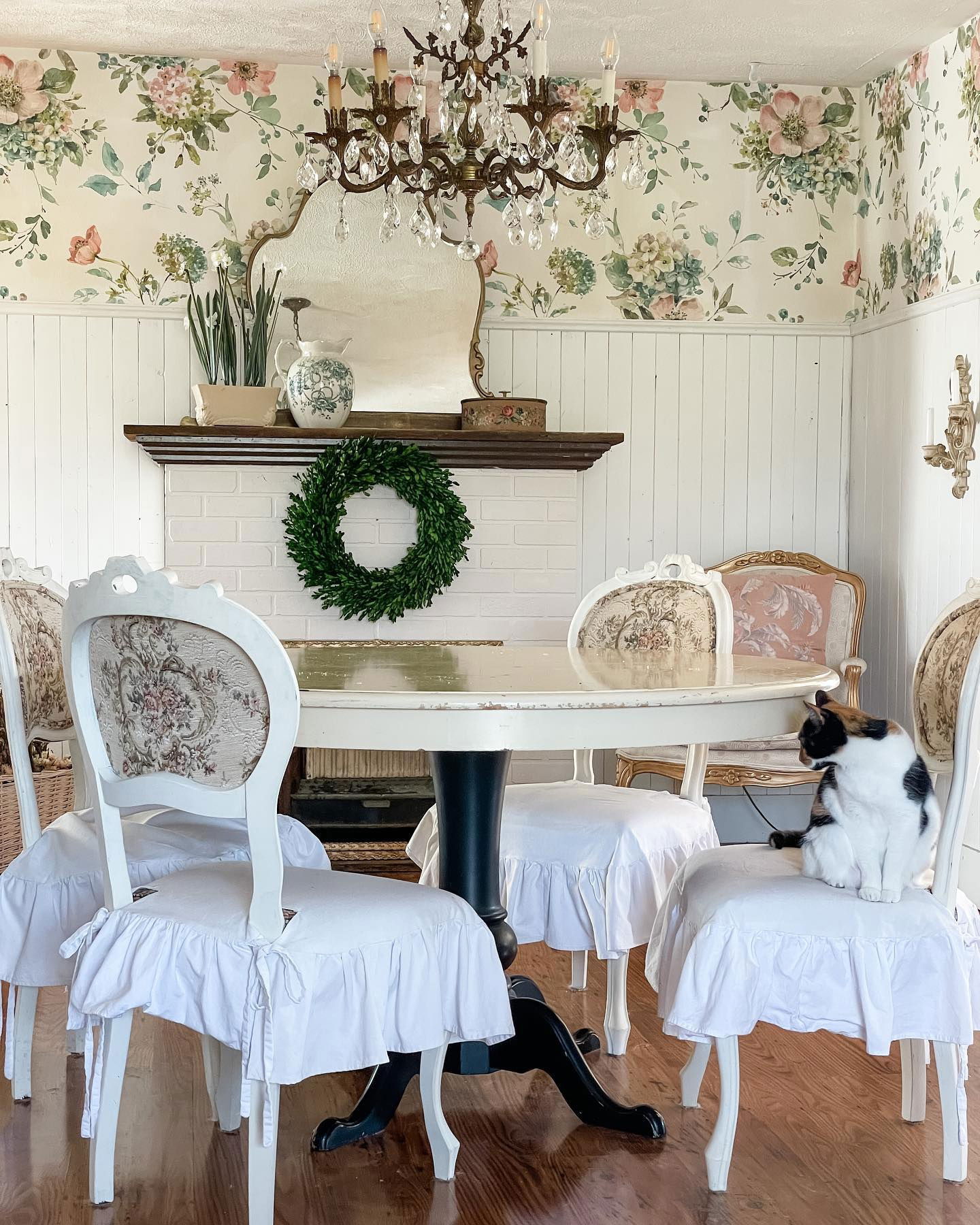 Shabby-Chic-Esszimmer mit floraler Tapete, weißen Stühlen mit Rüschenbezügen und einer schwarz-weißen Katze.