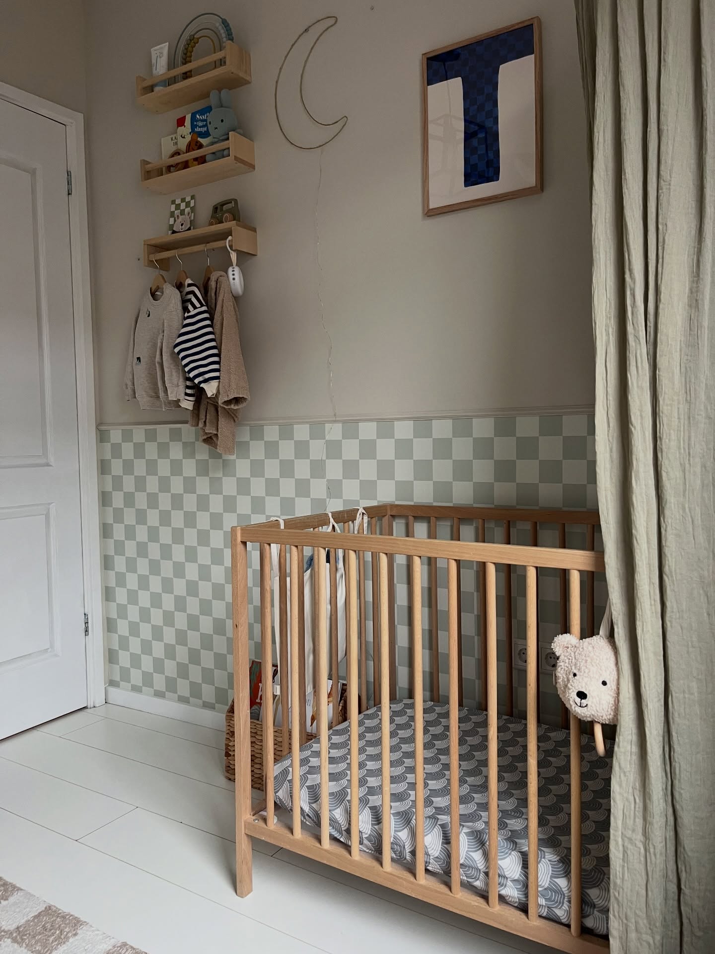 Wooden crib with a teddy bear on the side, a checkered wallpaper, and clothes hanging on a rack.