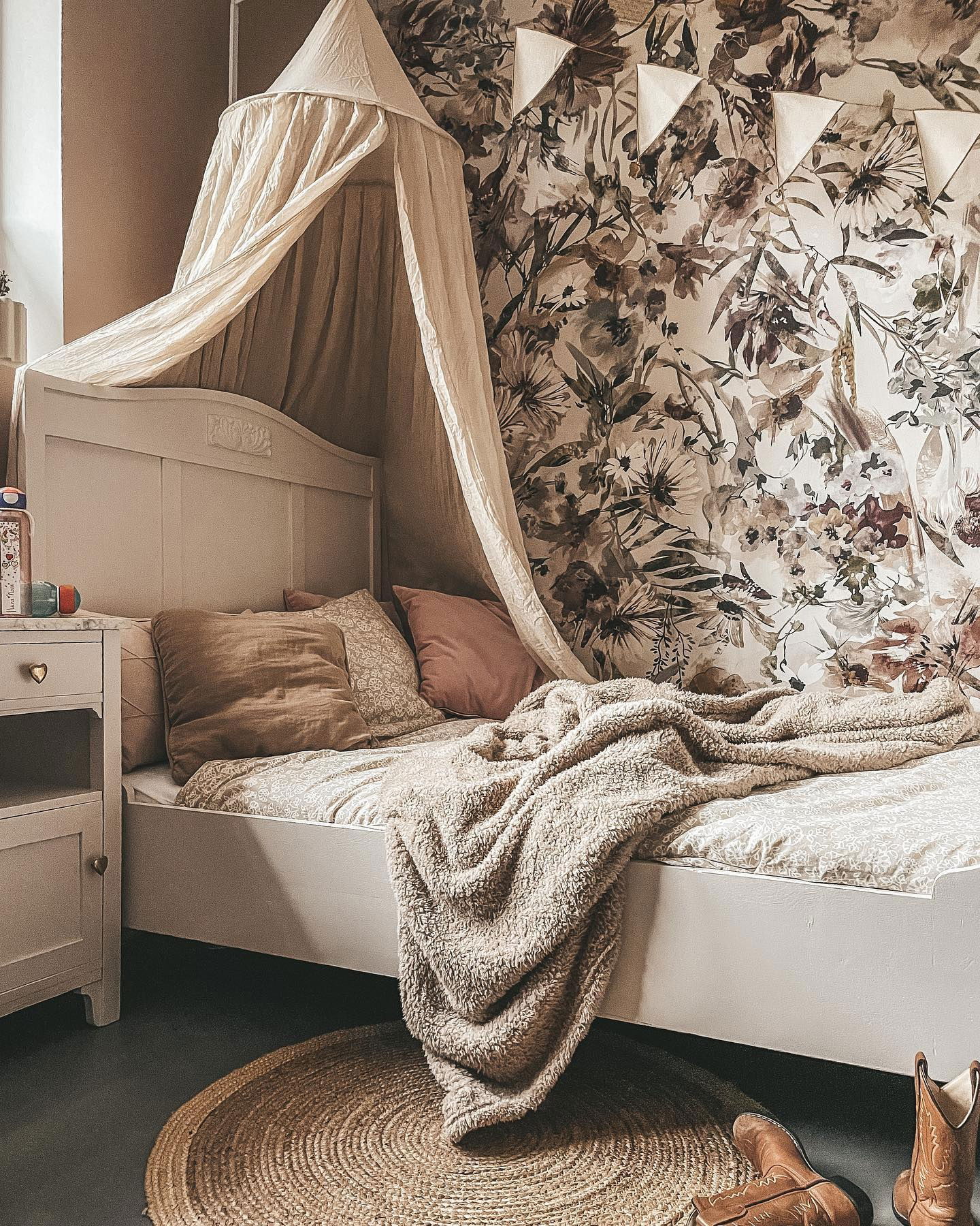 Cozy bedroom with white bed, canopy, floral wallpaper, and a round jute rug. Cowboy boots sit on the floor.