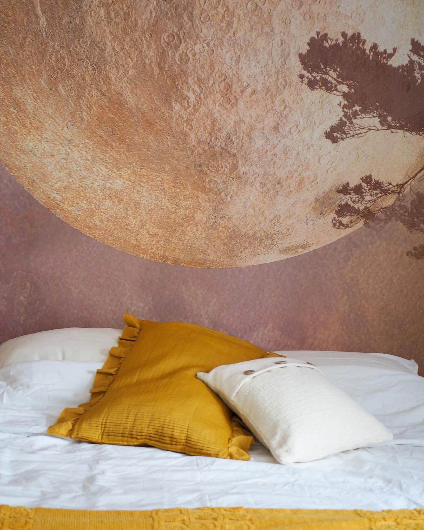 Schlafzimmer mit Wandbild des Mondes, weißen Laken und gelben Kissen.
