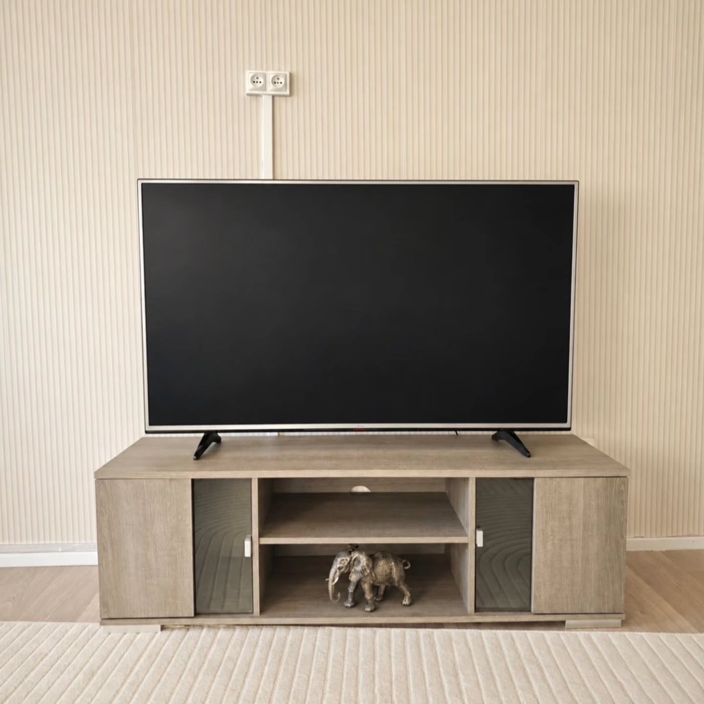 Gran televisor negro de pantalla plana sobre un soporte de entretenimiento de madera clara con una pequeña figura de elefante gris. Papel pintado de rayas claras y una alfombra.