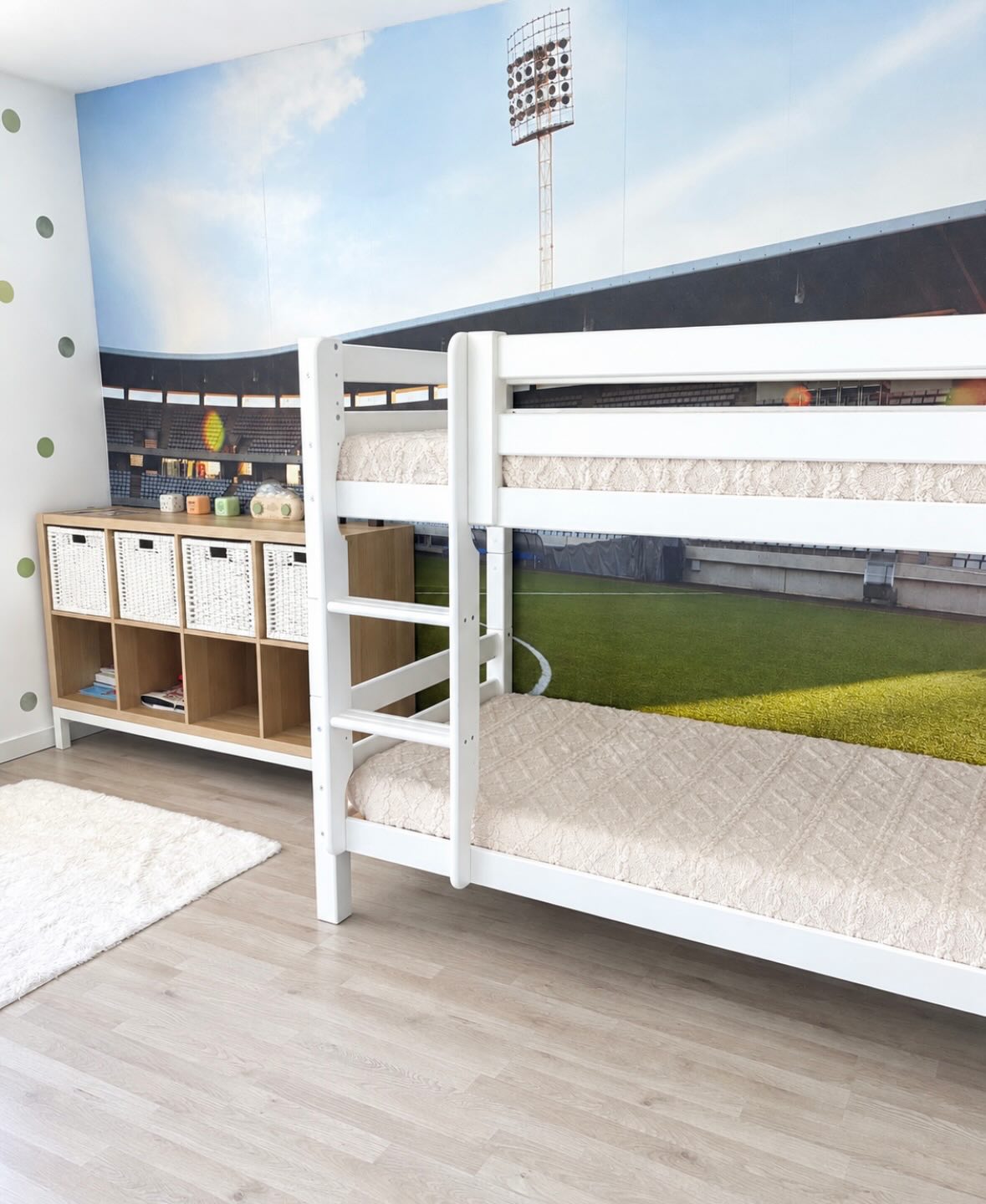 White wooden bunk beds with cream bedding against a stadium wall mural featuring a green field and stands. A light wood shelving unit with white baskets and green polka dots on the wall.
