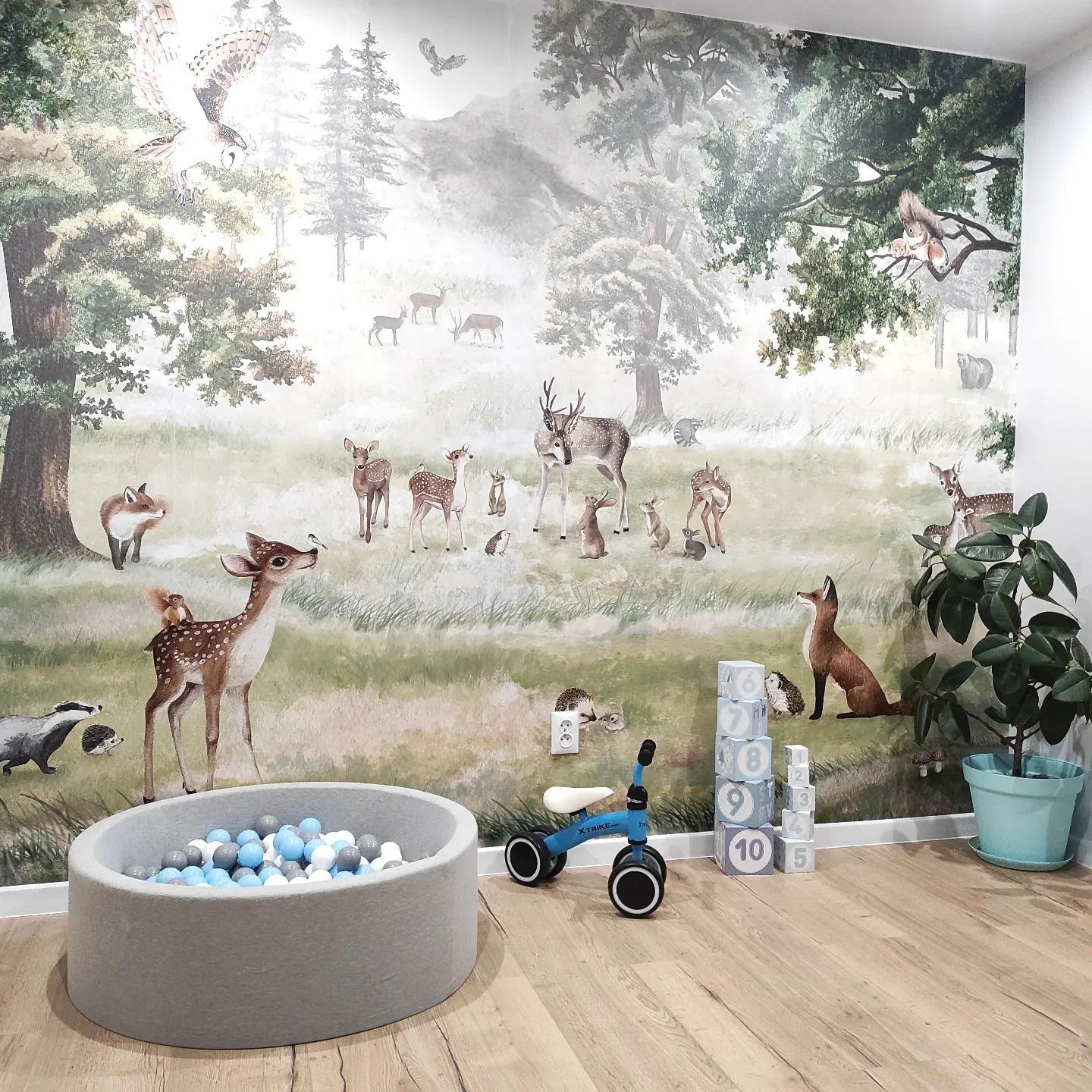 Chambre d'enfant avec papier peint sur le thème de la forêt, piscine à balles grise, draisienne bleue et blocs numérotés.