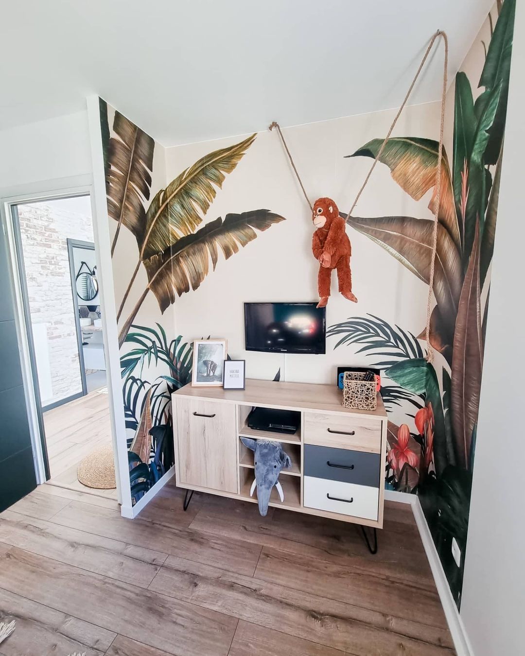 Living room with tropical wallpaper, light wood floors, and a wood media cabinet. A stuffed monkey hangs from the ceiling.