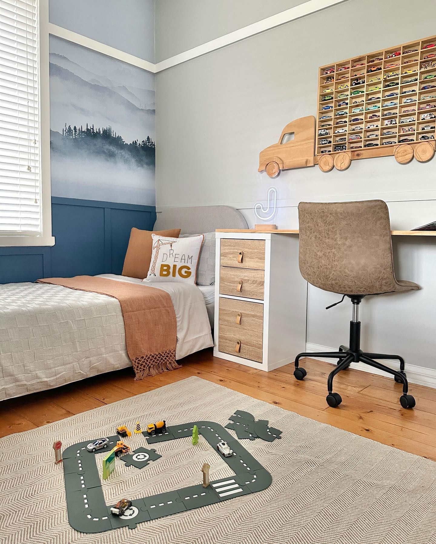 Child's bedroom with bed, desk, and toy car track on a herringbone rug. Wall art includes mountain mural and wooden truck-shaped car display.