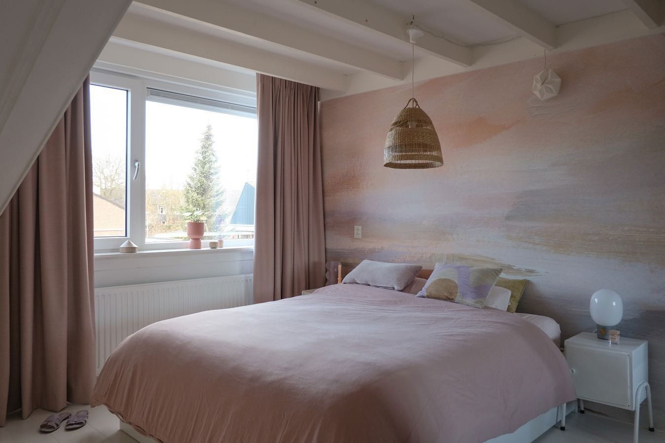 Soveværelse med pink sengetøj, vævet pendellampe og vægmaleri. Vinduesudsigt til træ.