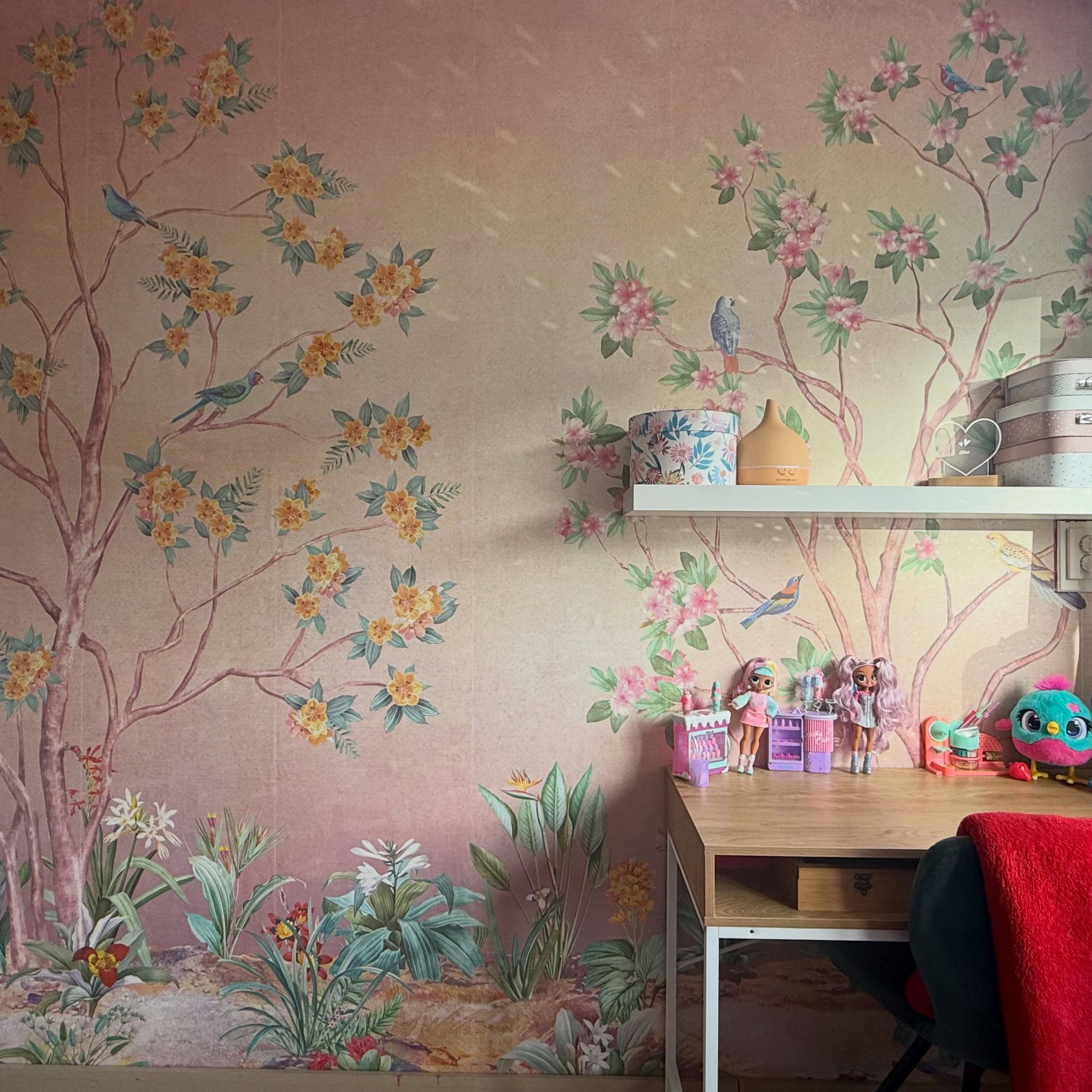 Wallpaper with painted trees, yellow and pink flowers, blue birds, and green foliage. A light wood desk with toys sits below a white shelf.