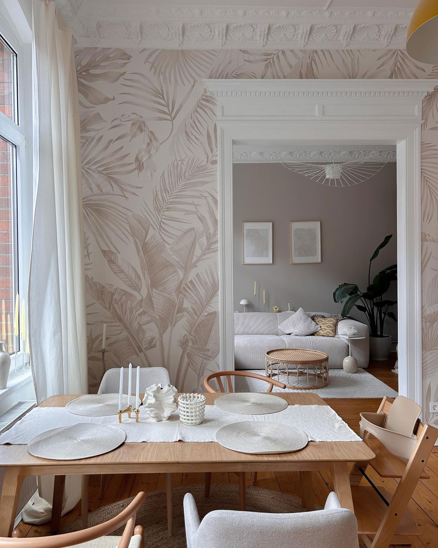 Dining room with tropical wallpaper, light wood table set for four, and view into a living room.