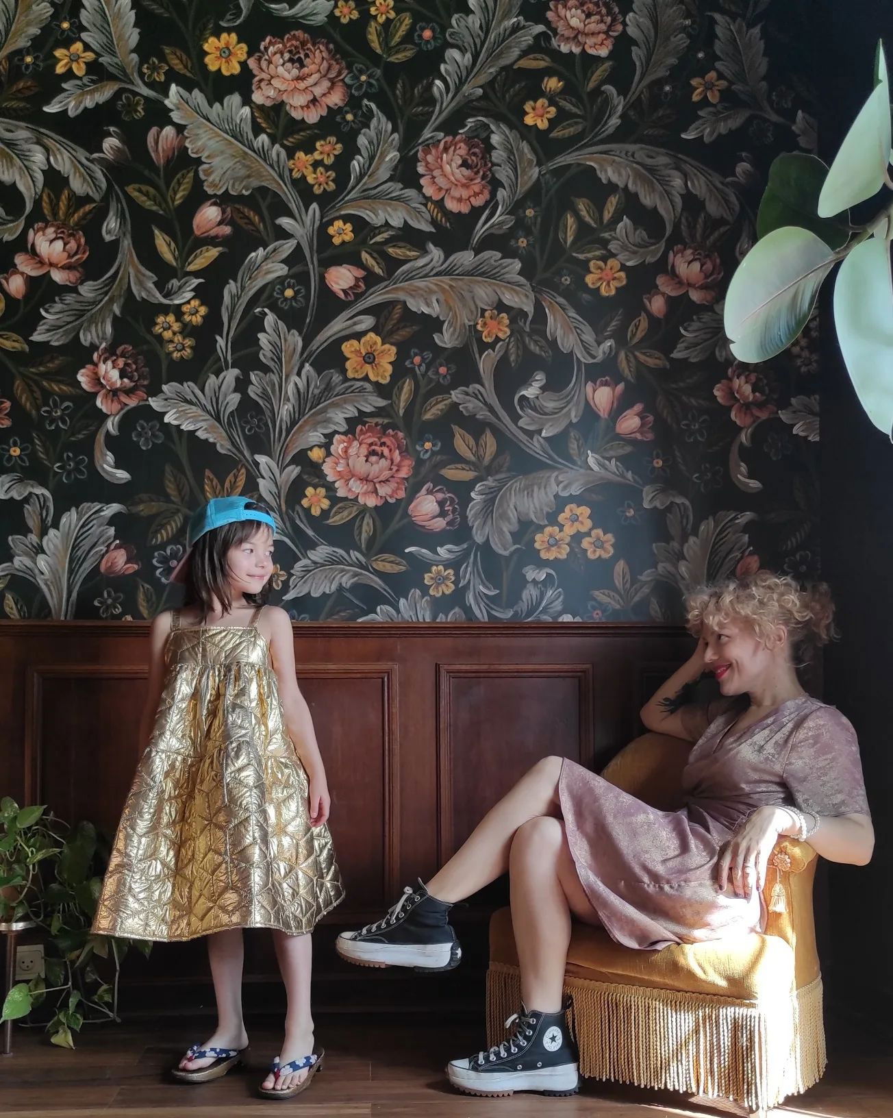 A girl in a gold dress and blue cap stands next to a woman in a pink dress and Converse sneakers. Floral wallpaper.