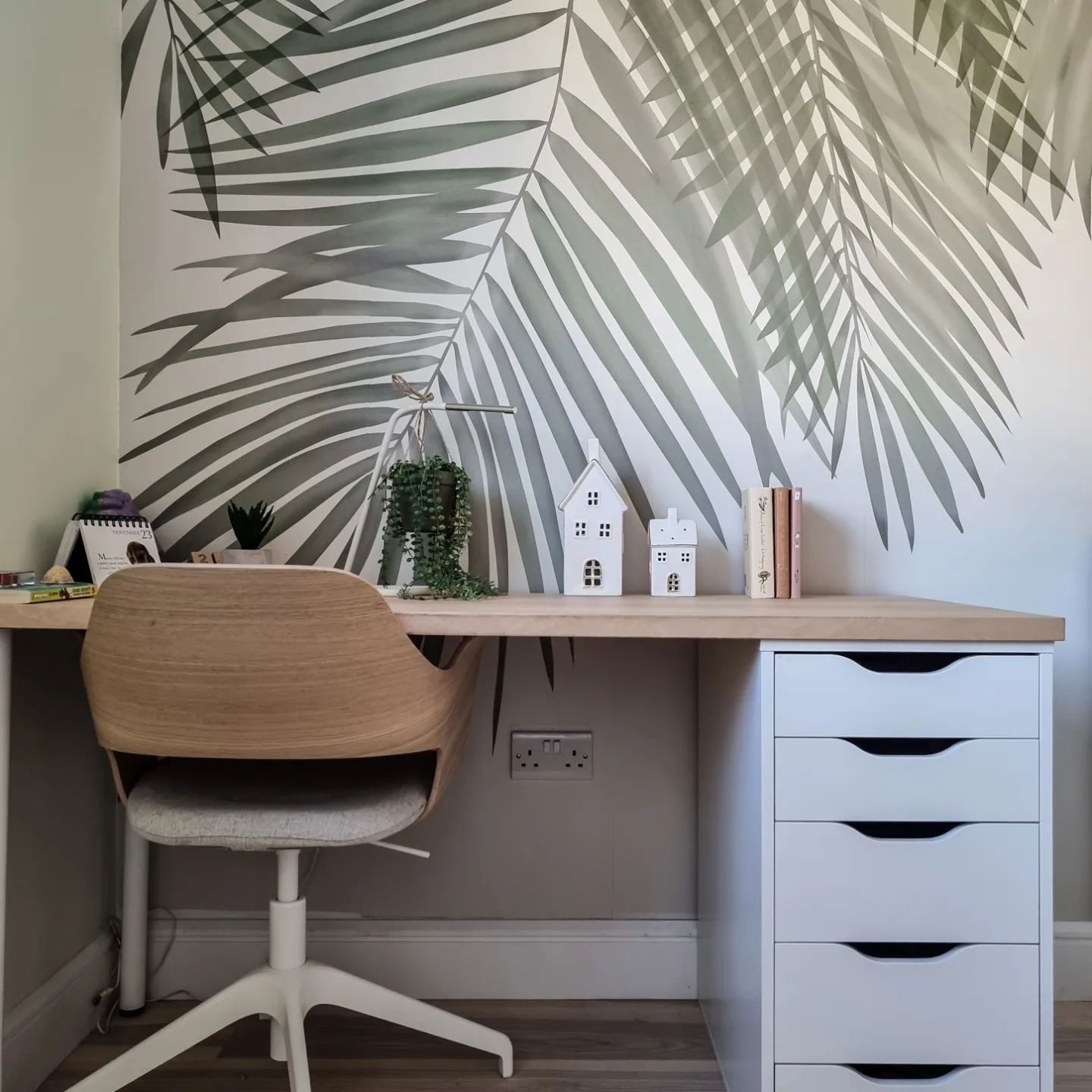 Home office setup with light wood desk, white drawers, and a wood and white swivel chair. Palm leaf wallpaper.