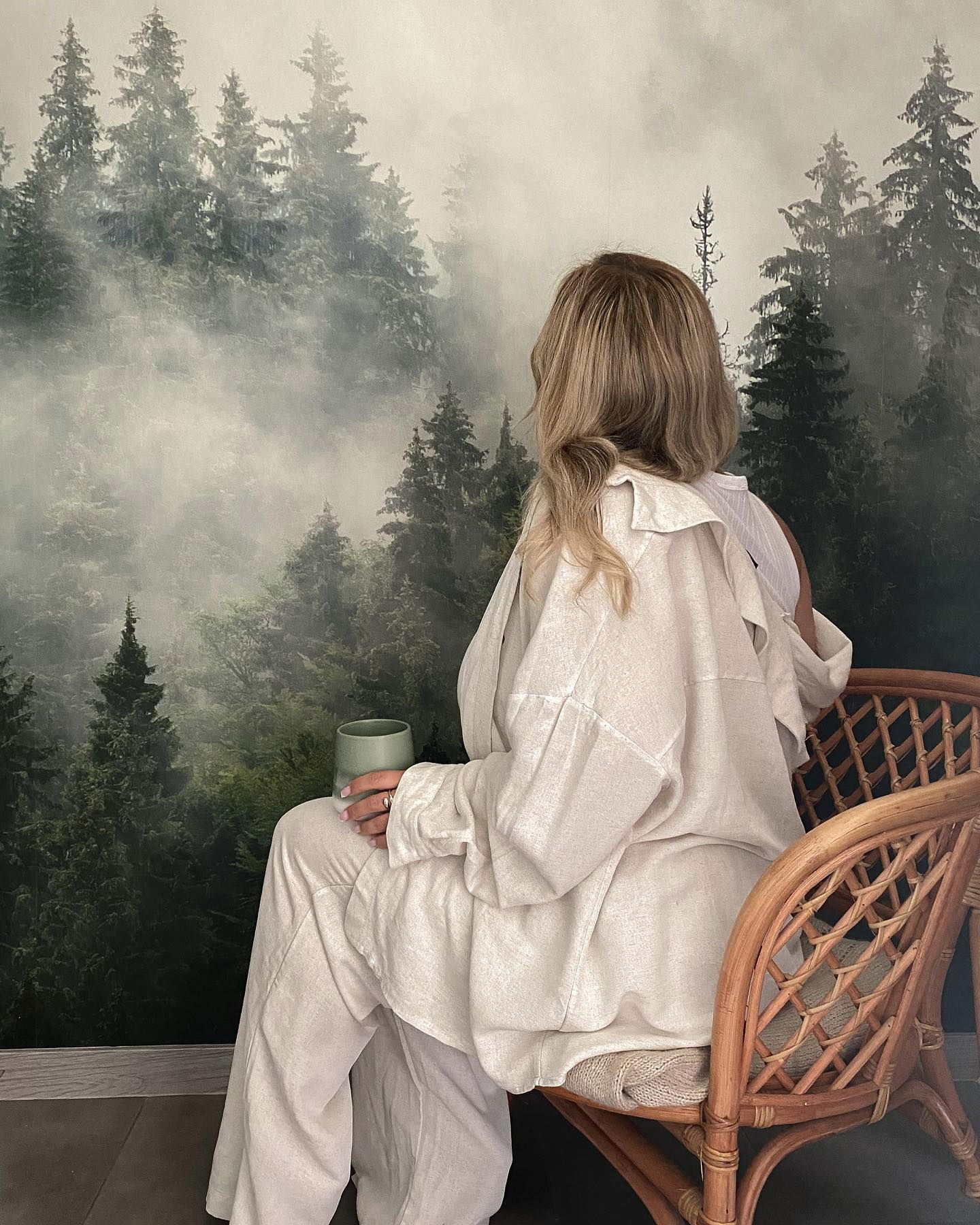 Frau im Leinenoutfit sitzt mit Tasse und blickt auf ein Waldwandbild. Korbstuhl.