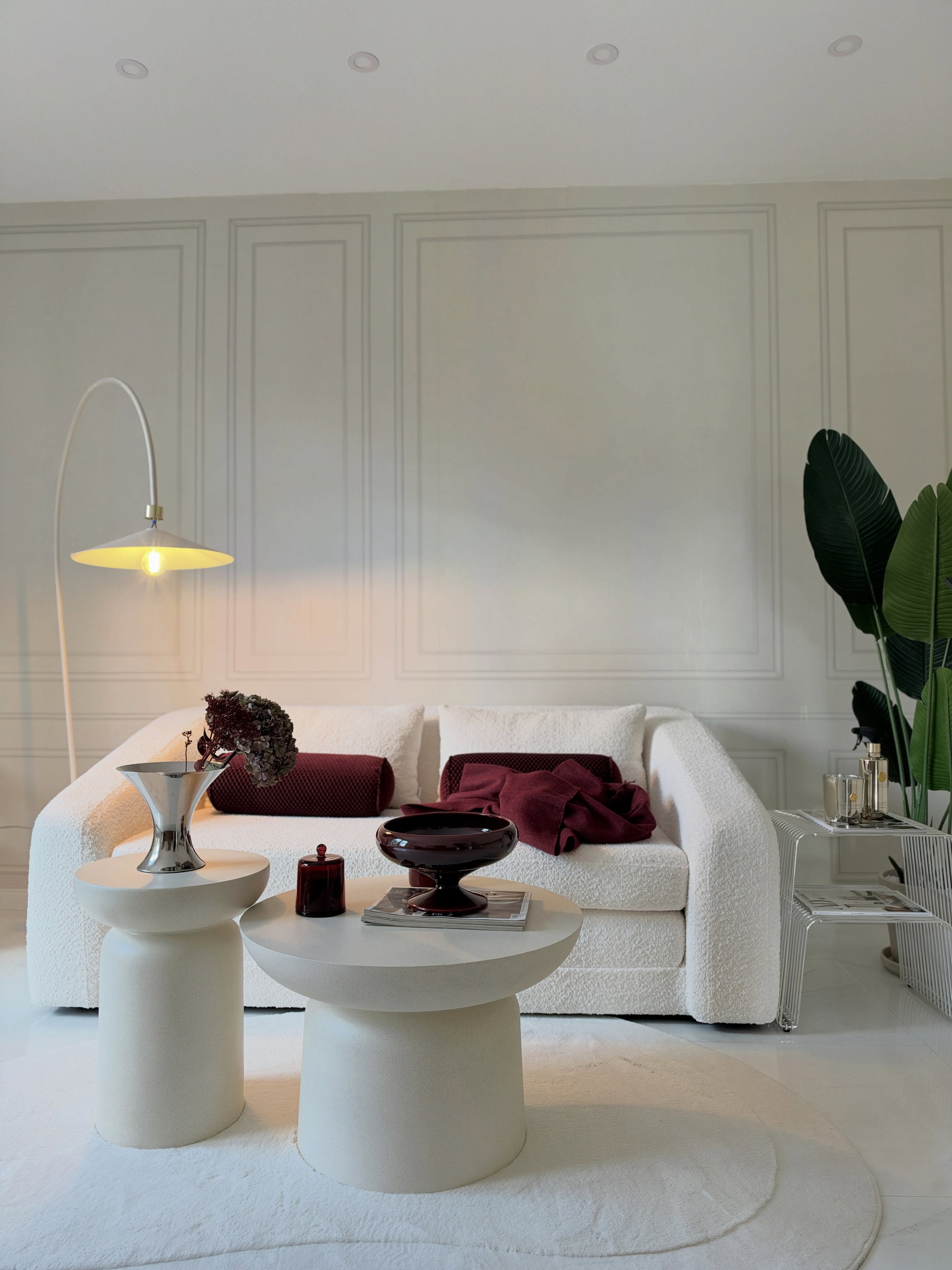 Un canapé texturé blanc avec des coussins et un jeté rouge foncé, deux tables d'appoint rondes crème, un lampadaire incurvé blanc et une plante verte.