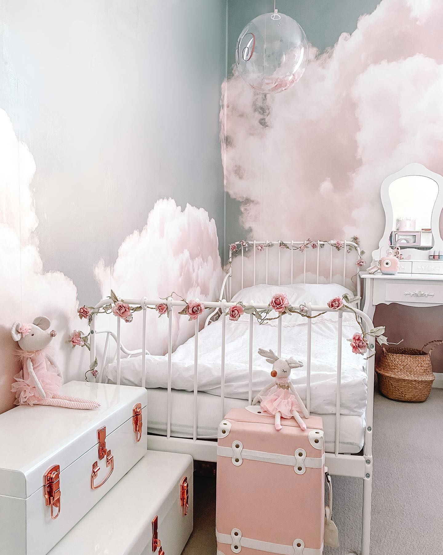 Child's bedroom with cloud wallpaper, white metal bed with rose garland, and pink and white storage trunks.
