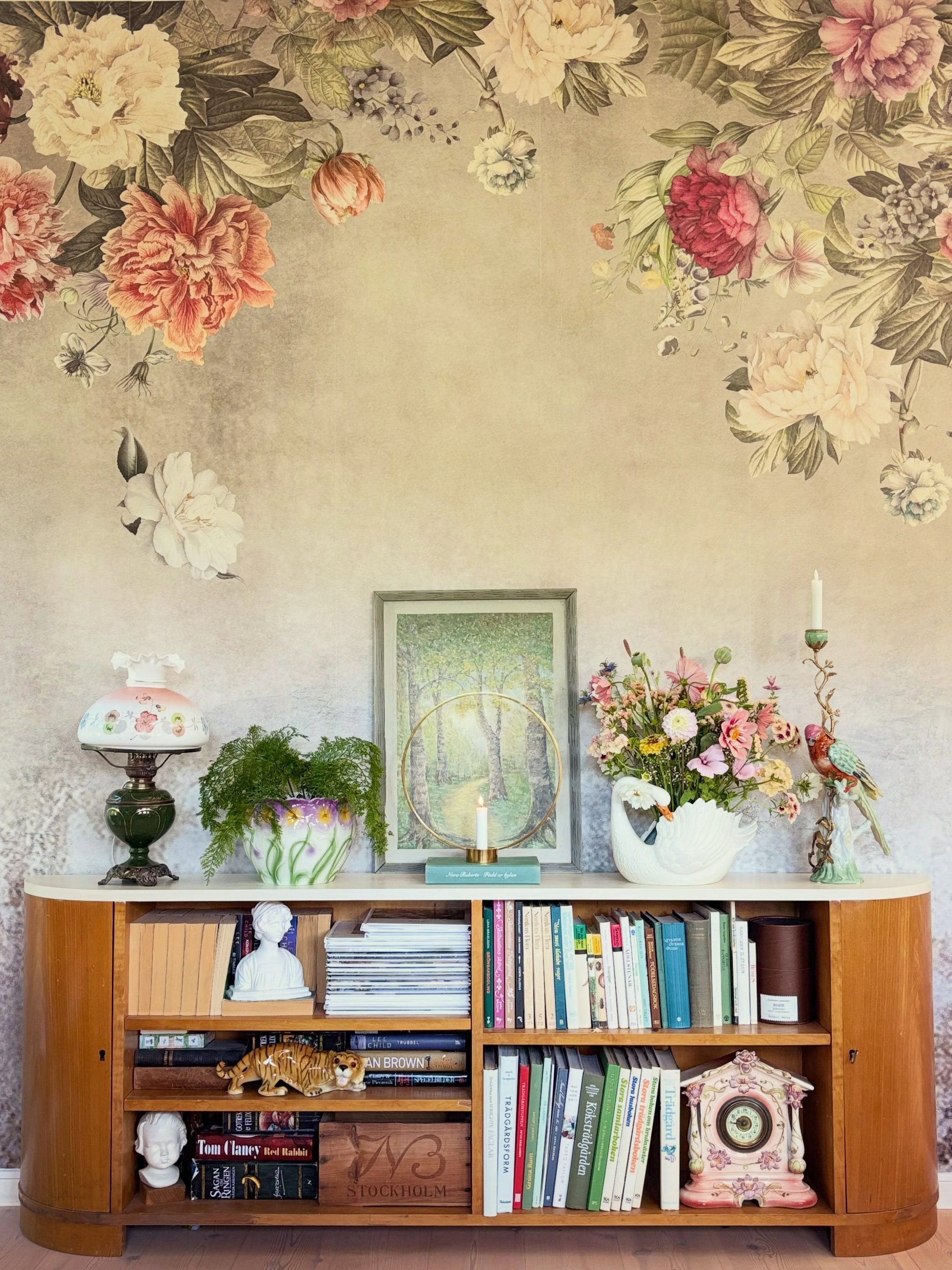 A light brown wooden cabinet with a white marble top, filled with books and decor, sits against a wall covered in large pink, red, and cream floral wallpaper. On top are a green lamp, potted fern, framed forest art, and a colorful flower bouquet in a swan vase.