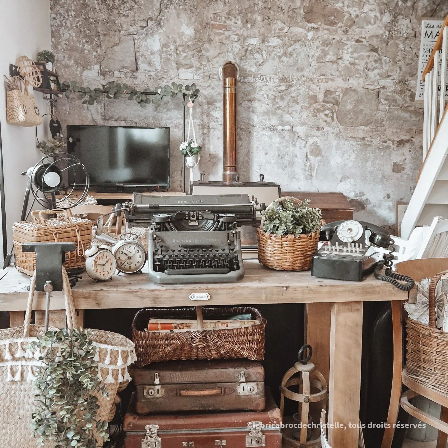 Vintage desk with typewriter, rotary phone, clocks, and stacked suitcases against a stone wall.