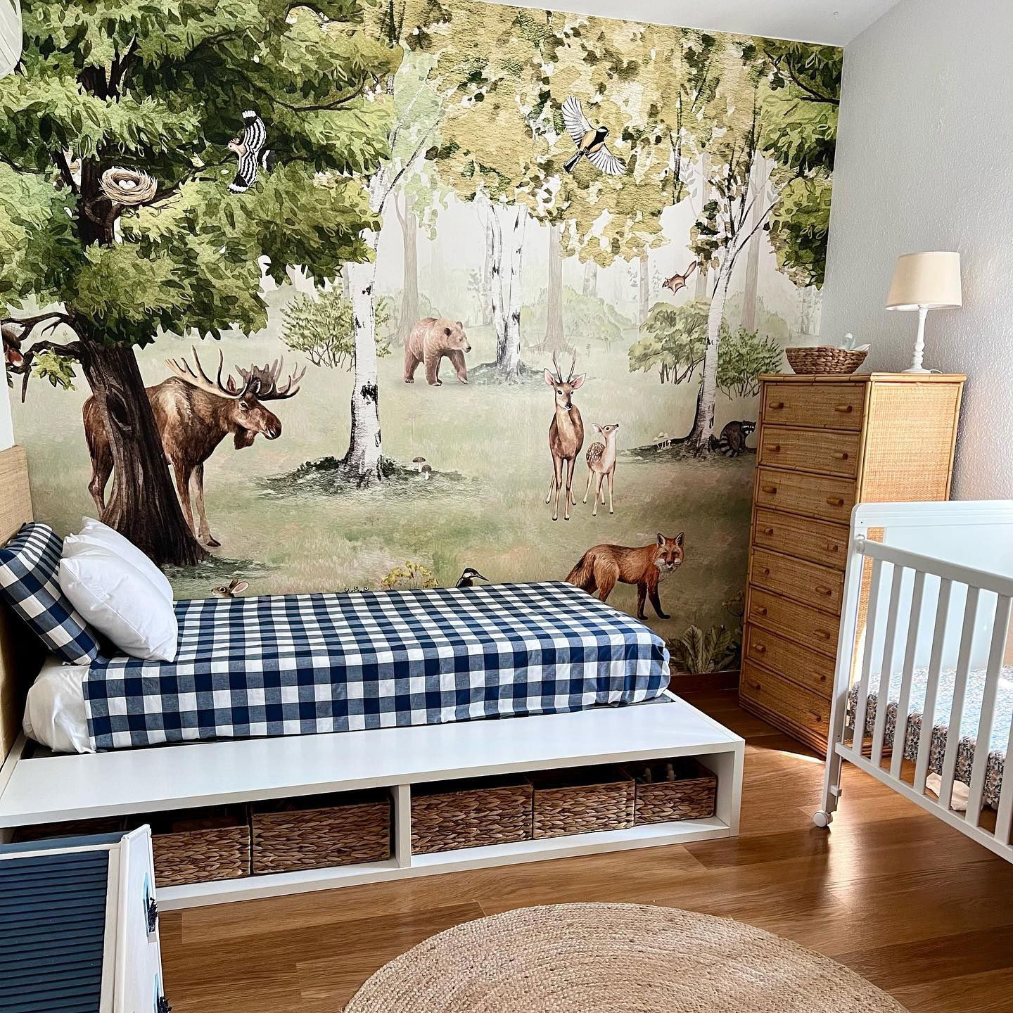 Stanza dei bambini con murale della foresta, culla bianca e letto a quadretti blu.