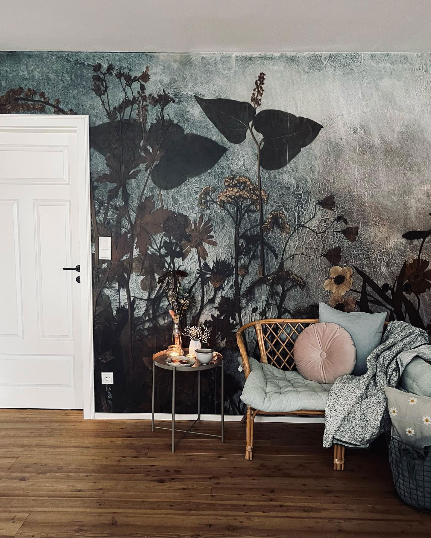 Hyggeligt hjørne af et værelse med blomstret tapet, rattan-stol med puder og et lille sidebord med et stearinlys.