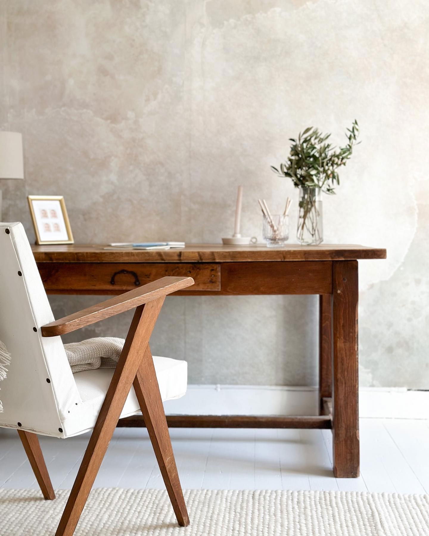 Interior con escritorio de madera, silla con cojín blanco y pared con textura neutra.