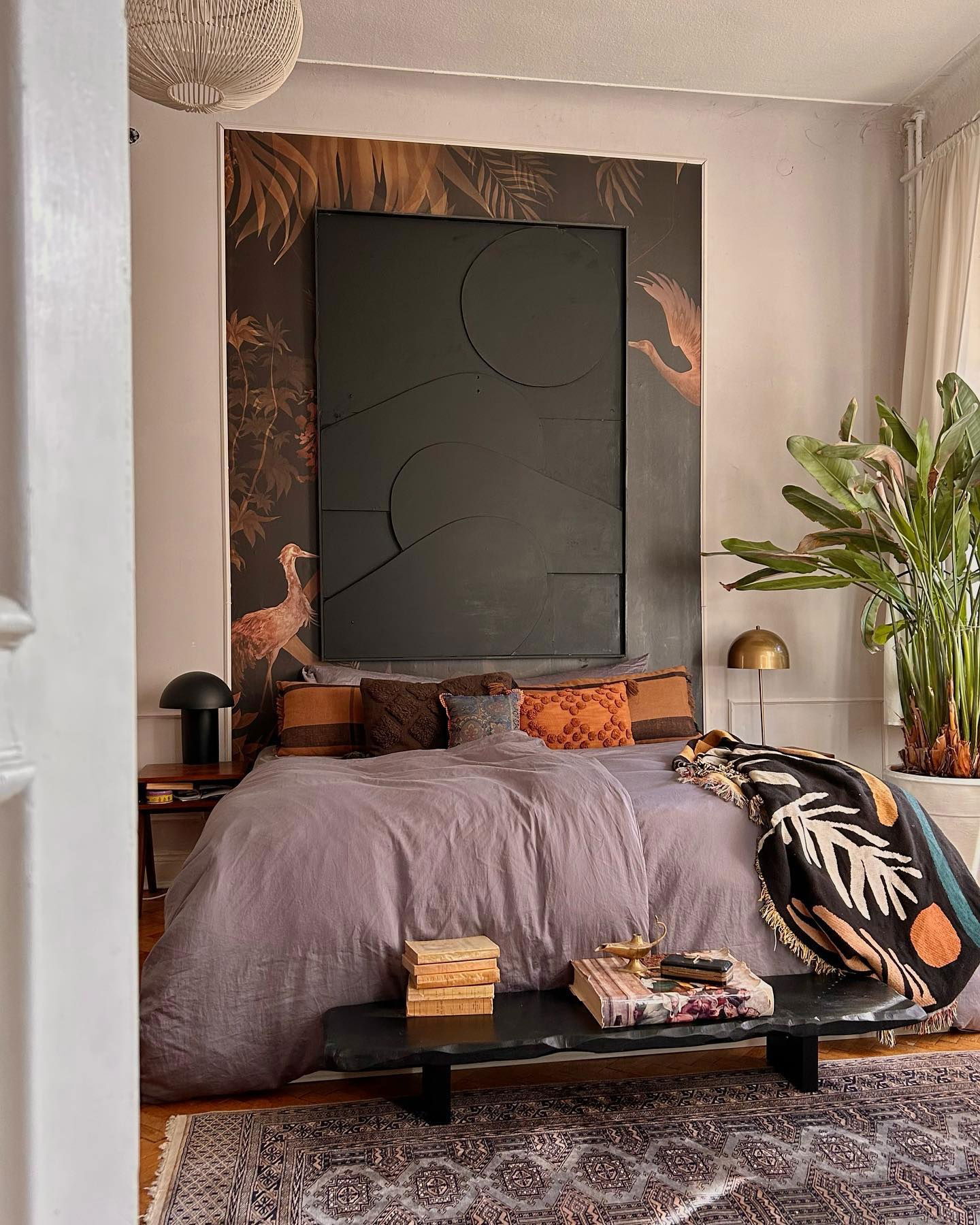 Schlafzimmer mit lila Bettwäsche, gemusterten Kissen und einer schwarzen Überwurfdecke. Eine schwarze Steinbank steht am Fußende des Bettes.